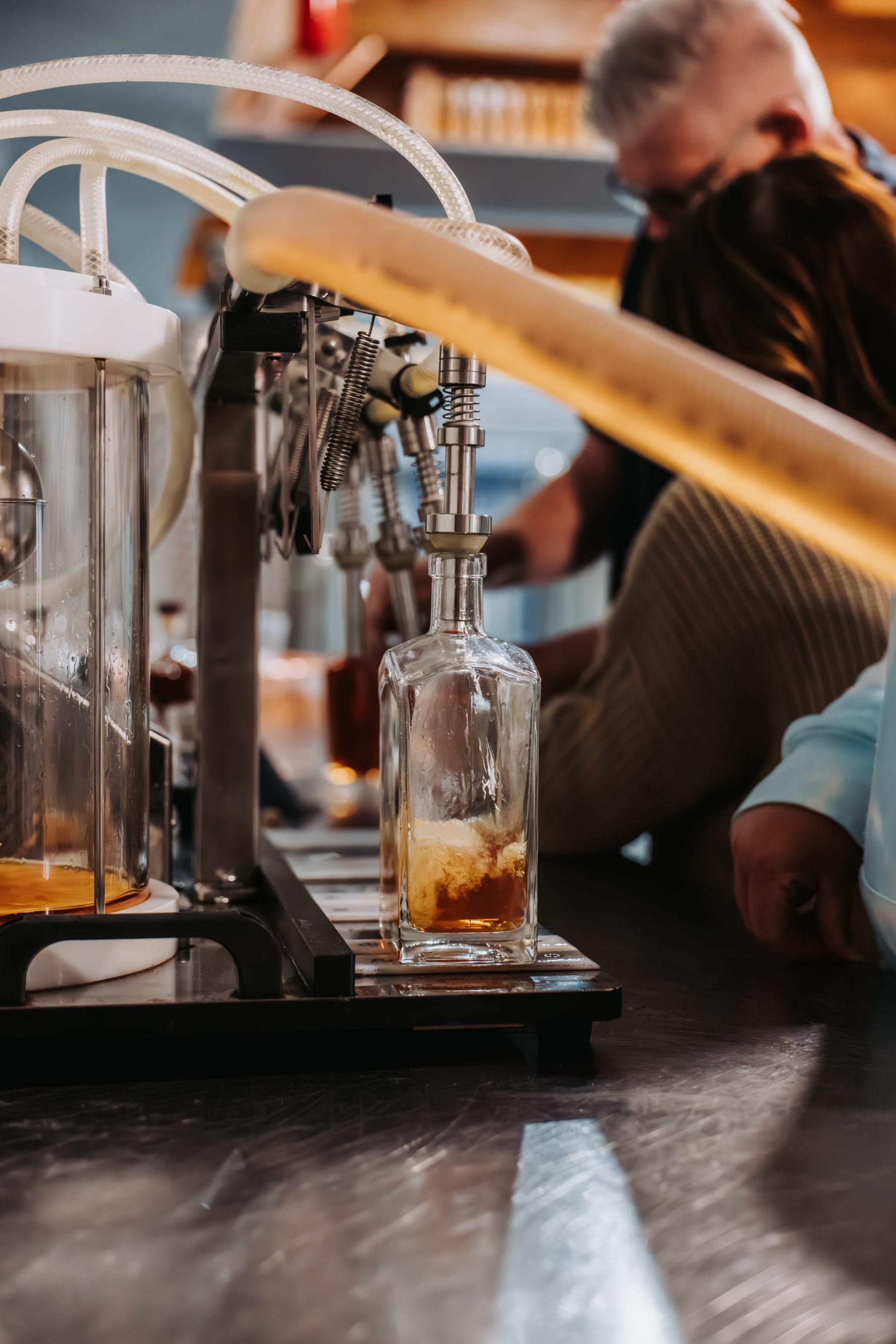 A bottle being filled with amber liquid from a machine, with a person in the background.