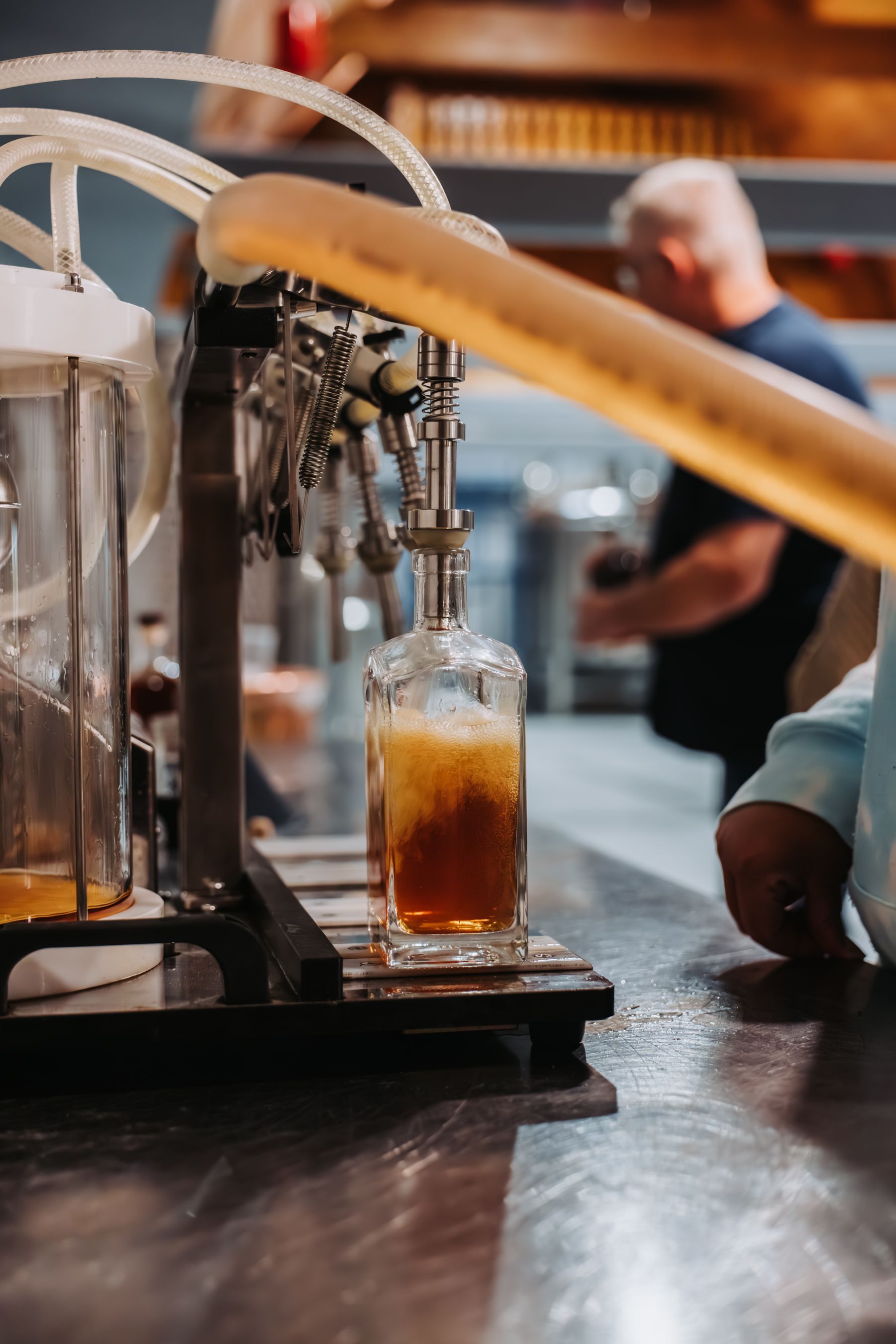 A bottle being filled with a dark liquid from a tap system; a person in the background.