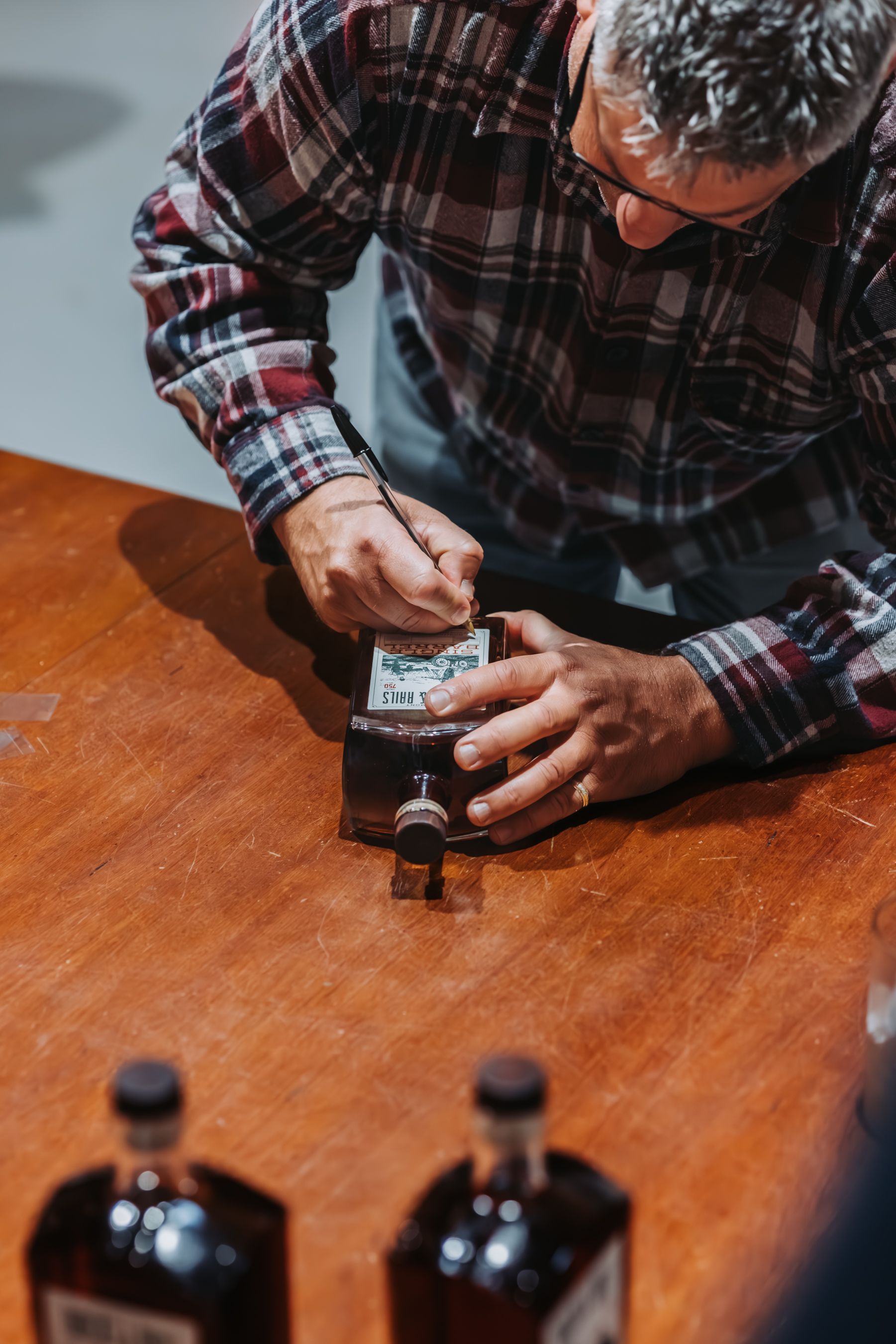 Man in plaid shirt labels a liquor bottle on a wooden table.