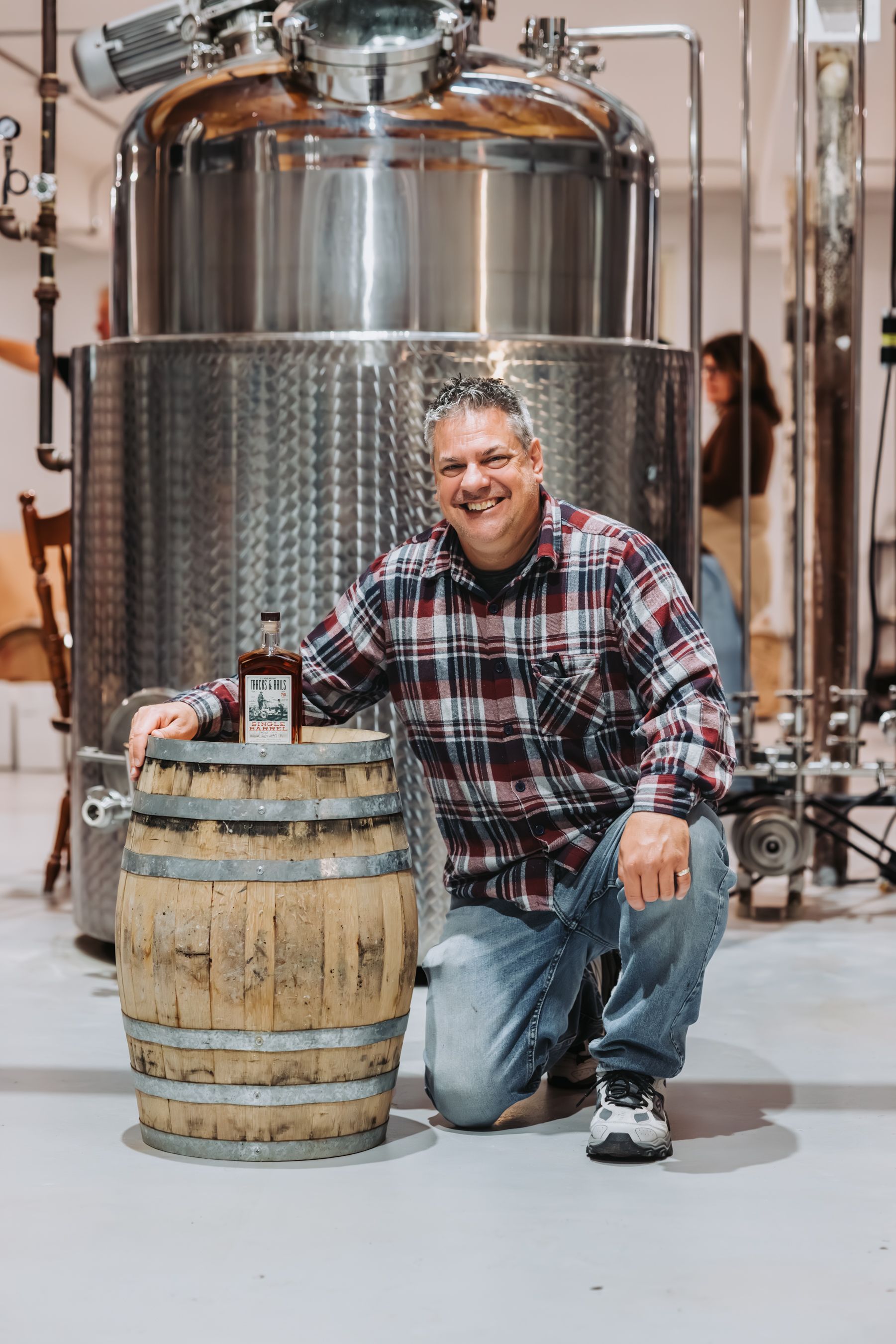 Man kneeling next to a barrel, product bottle on top, in a distillery with stainless steel tanks.