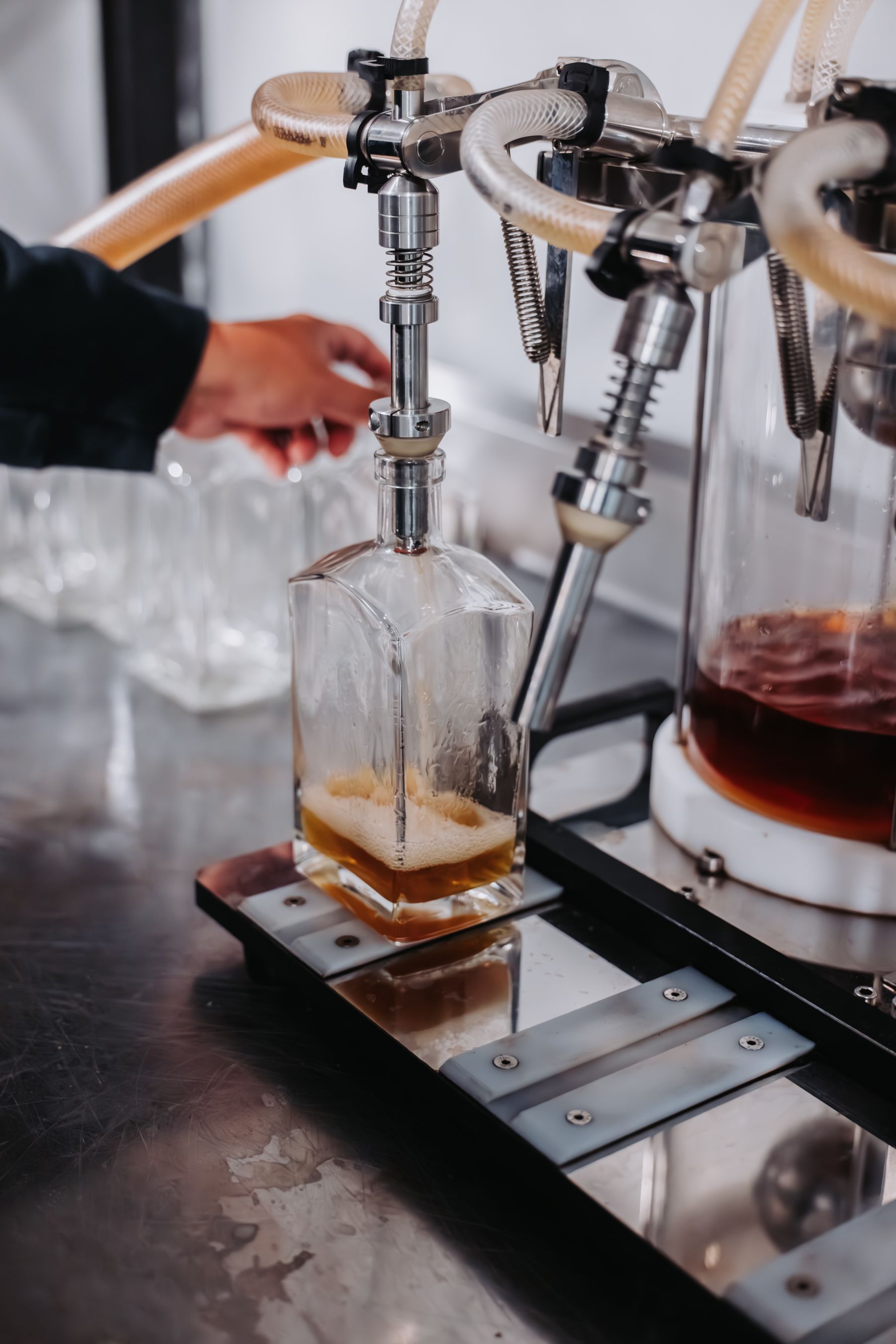 Man pouring liquid from a machine into a glass bottle. Beige liquid, stainless steel, clear bottles.