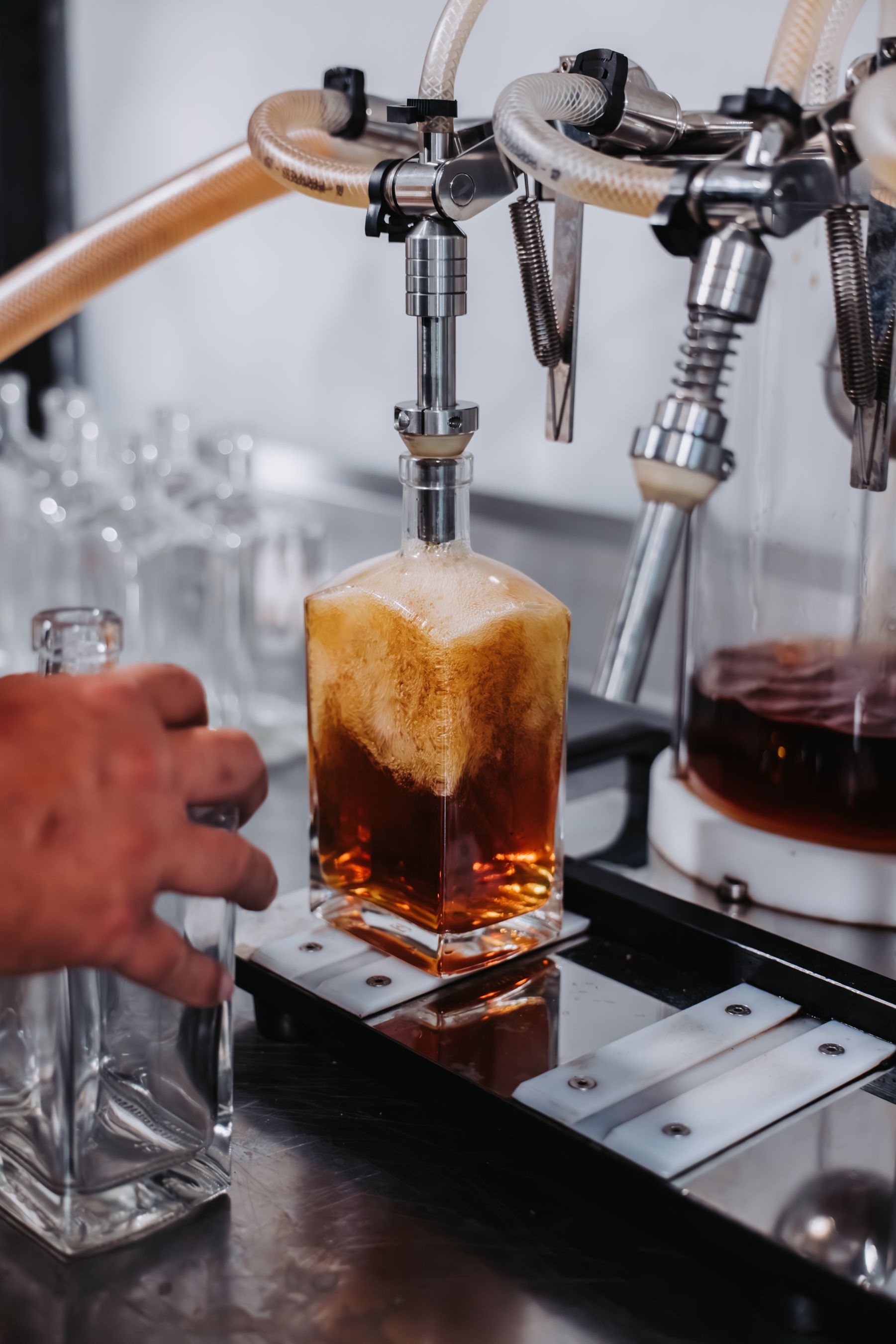Bottling machine filling a square glass bottle with amber liquid; a person's hand adjusts a bottle on a metal track.