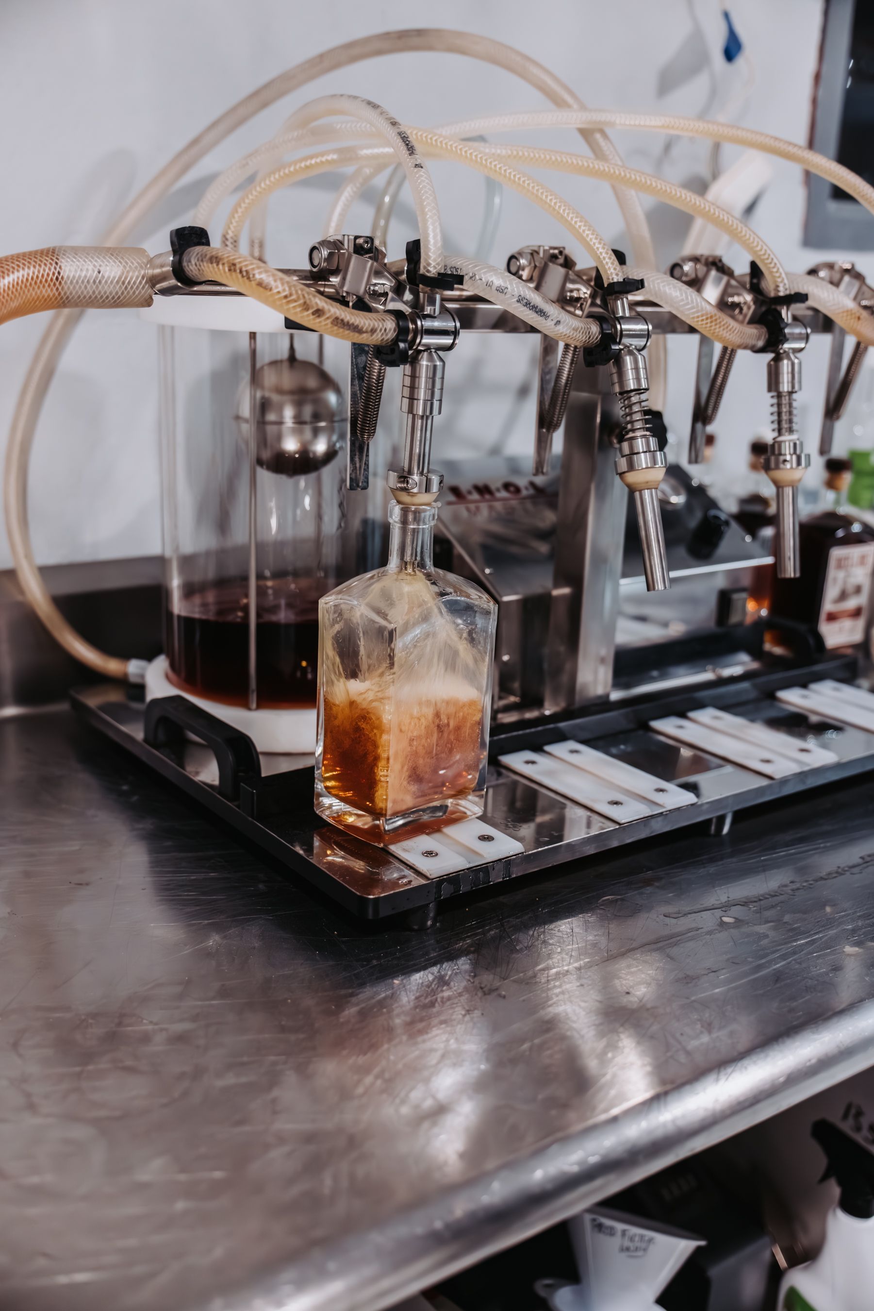 A drink dispenser pouring dark liquid into a glass bottle. Stainless steel taps and clear tubing.