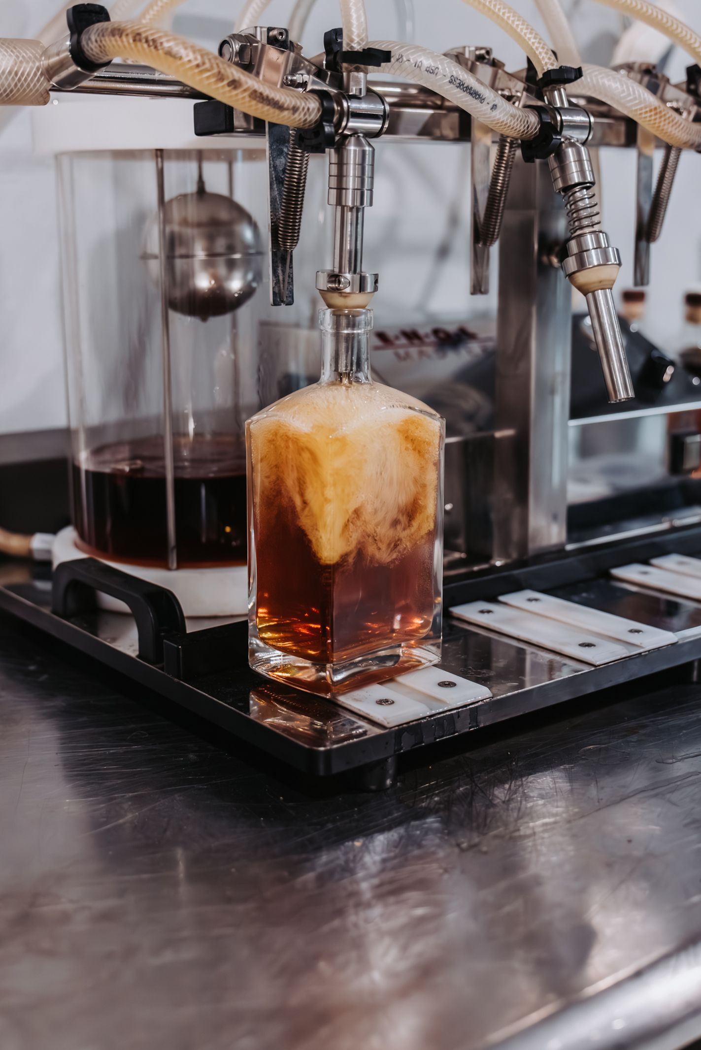 A decanter being filled with a dark liquid by a machine, generating foam. Stainless steel counter in a bar setting.