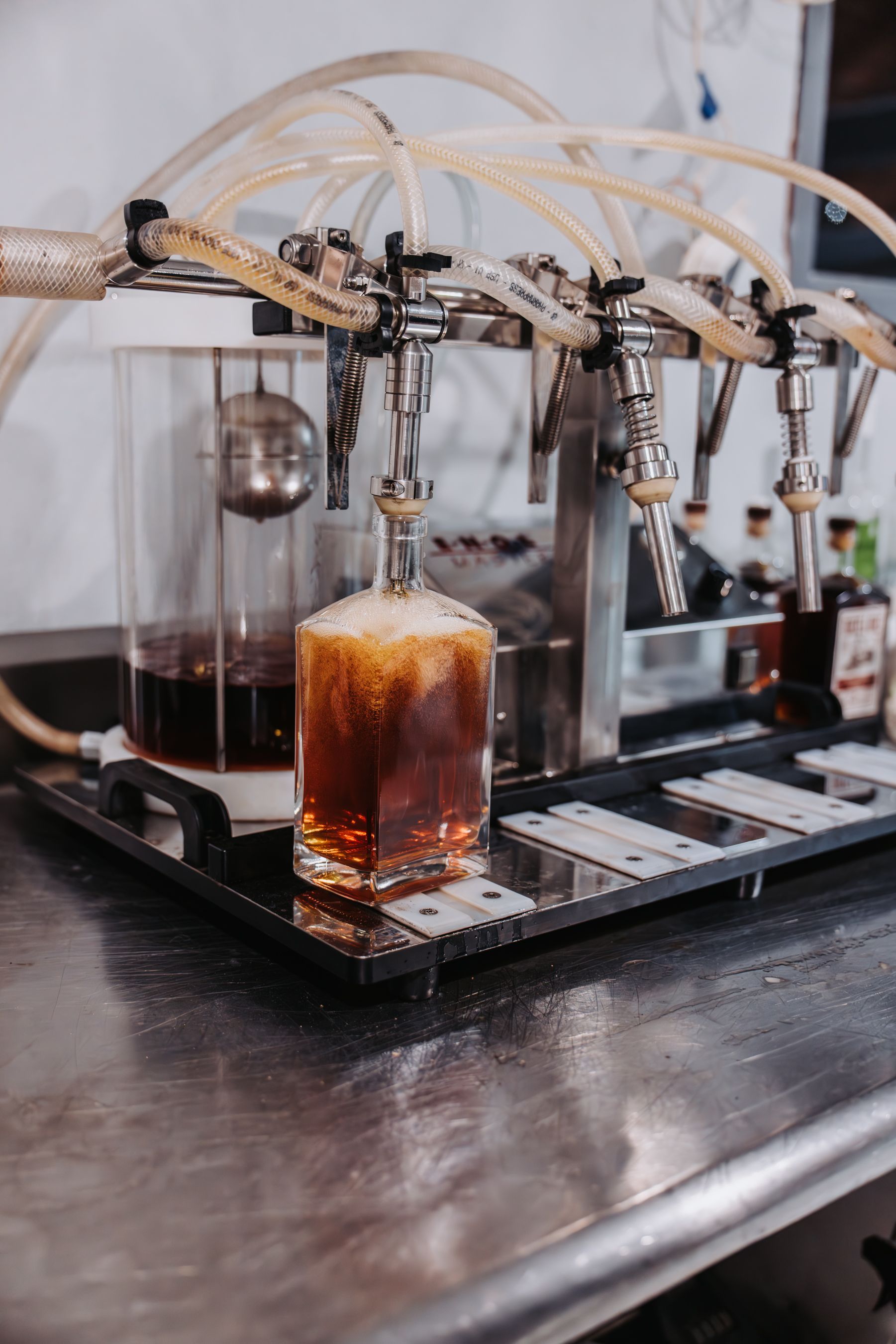 A bottle being filled with a dark liquid from a draft system with multiple taps, on a stainless steel surface.