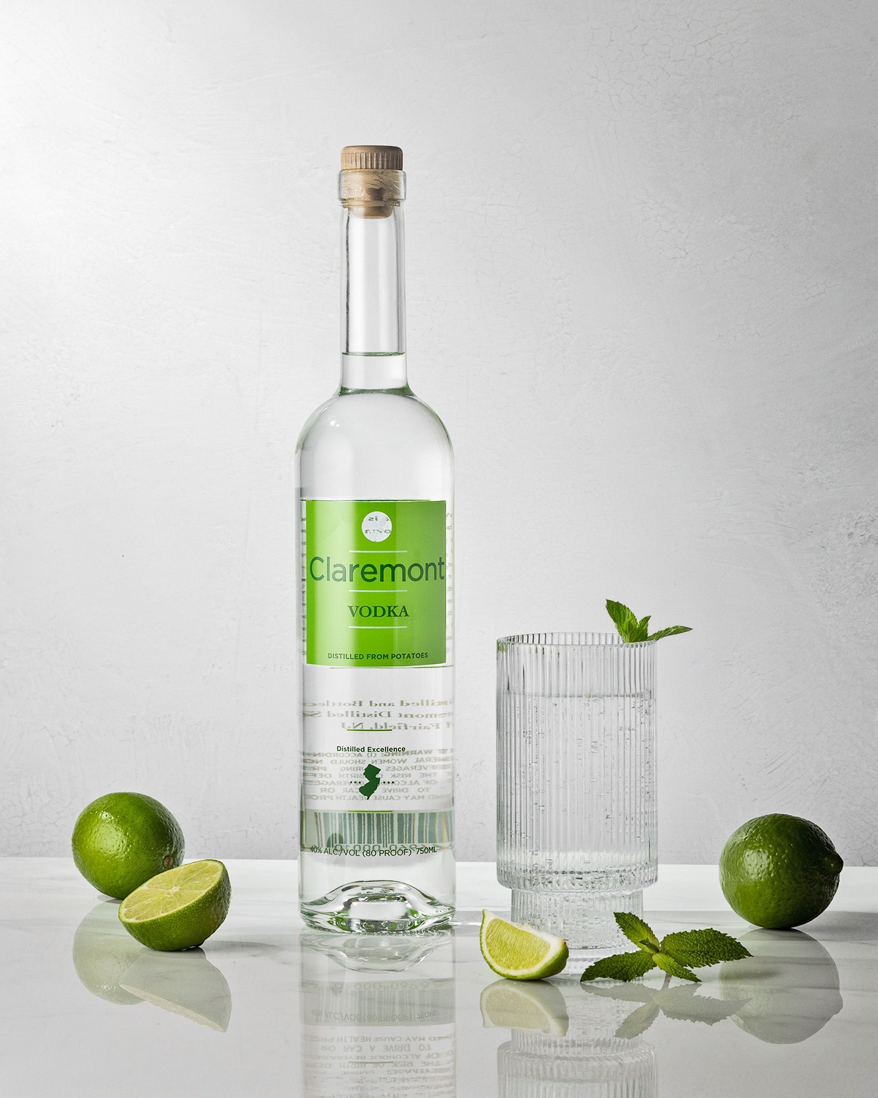 Bottle of clear liquid with a green label, surrounded by limes, mint, and a glass of beverage.