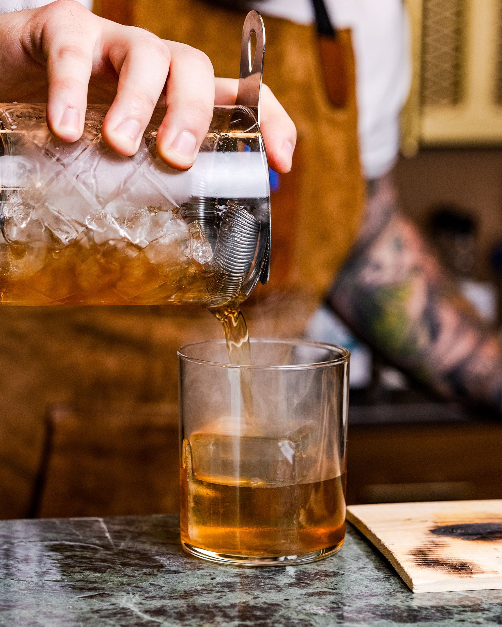 Bartender pouring a brown cocktail from a shaker into a glass with ice.