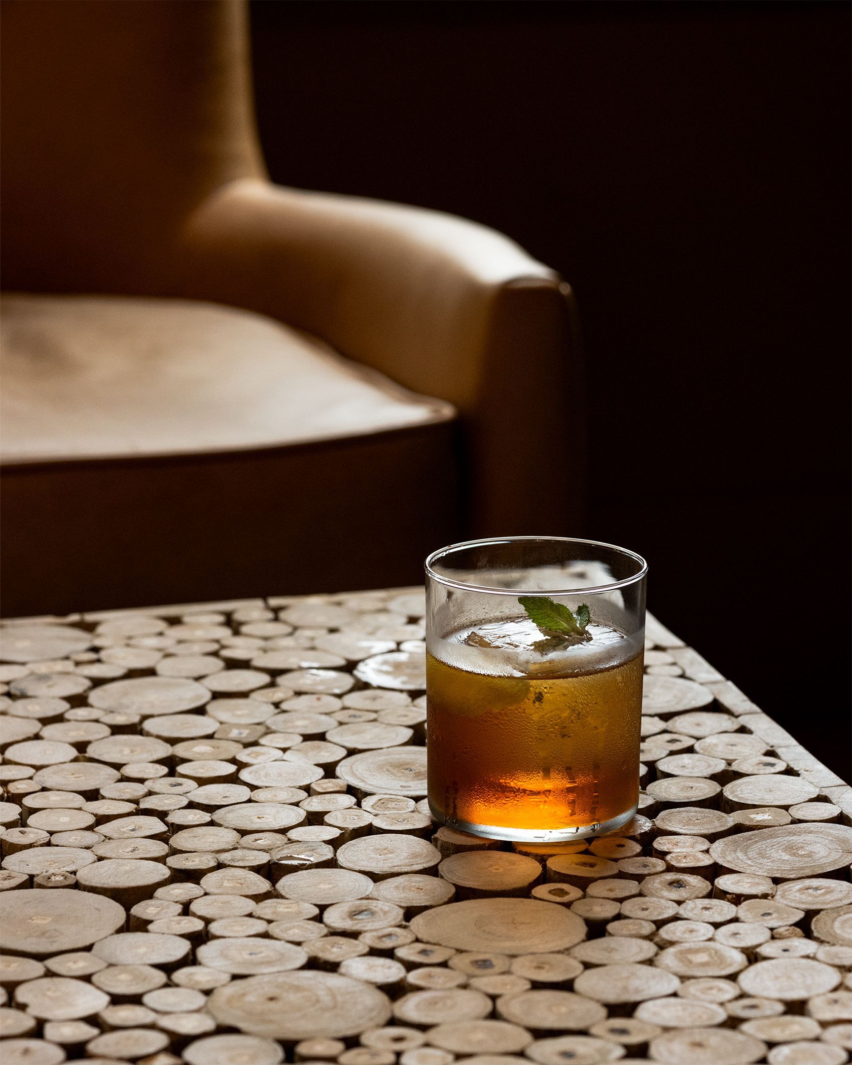 Cocktail in a glass with mint garnish on a wood-mosaic table, an armchair in the background.