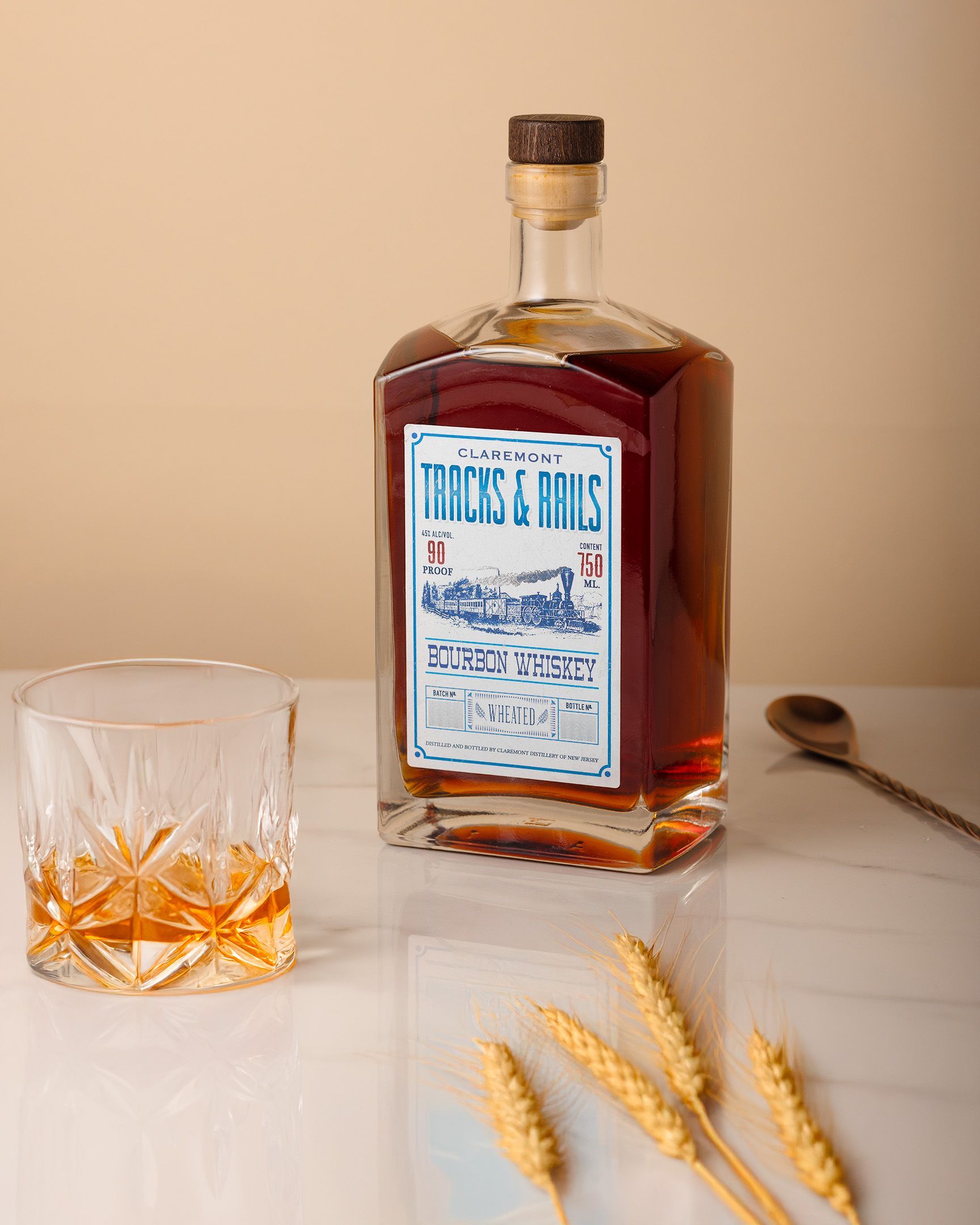 Bottle of whiskey with a glass, spoon, and wheat on a white surface, against a tan backdrop.