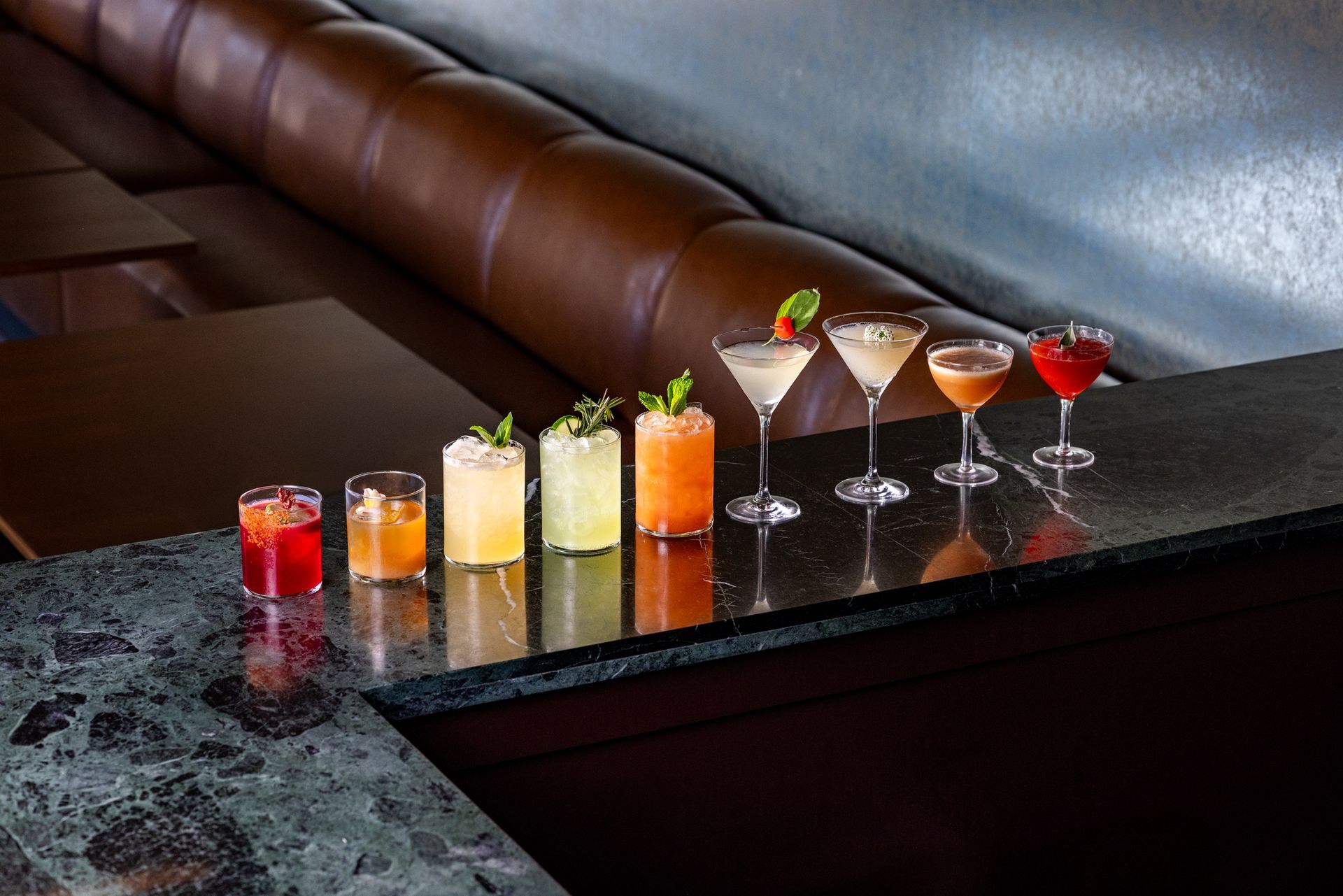 Row of colorful cocktails on a bar, with a dark brown booth in the background.