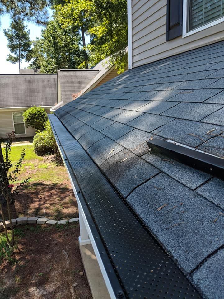 A view along a grey shingled roofline, featuring a black metal gutter guard, extending toward a house and yard.