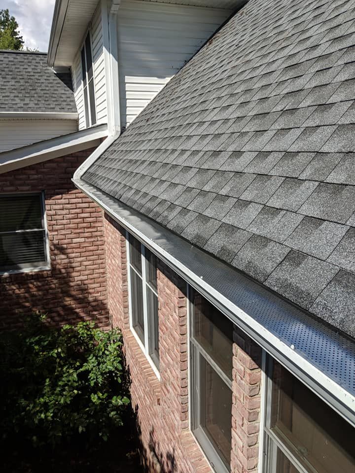 A high-angle view of a brick house exterior showing grey asphalt shingles, white siding, and a gutter with a metal guard.