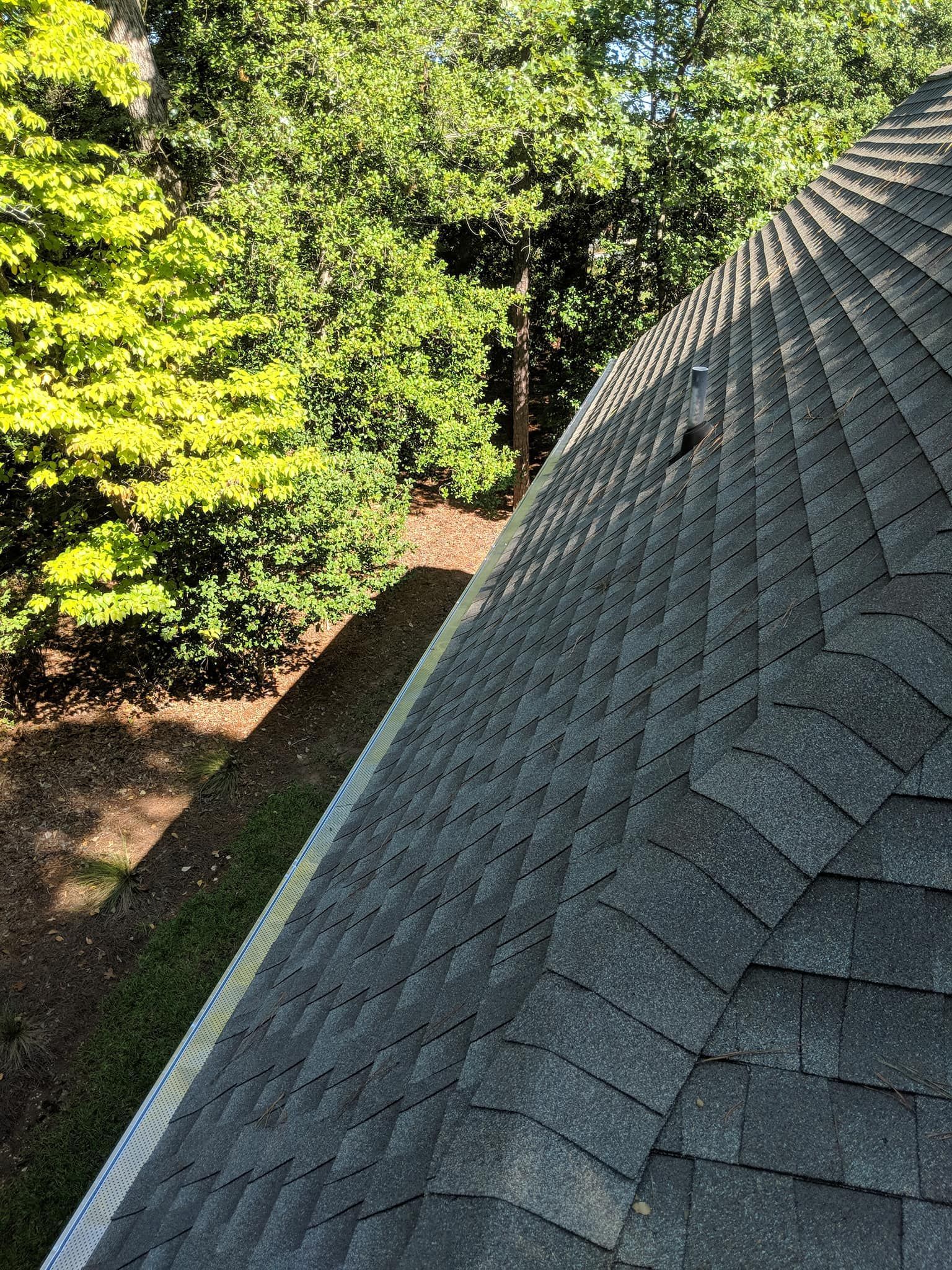 A high-angle view looking down at a gray shingled roof next to a lush green tree and a patch of ground.
