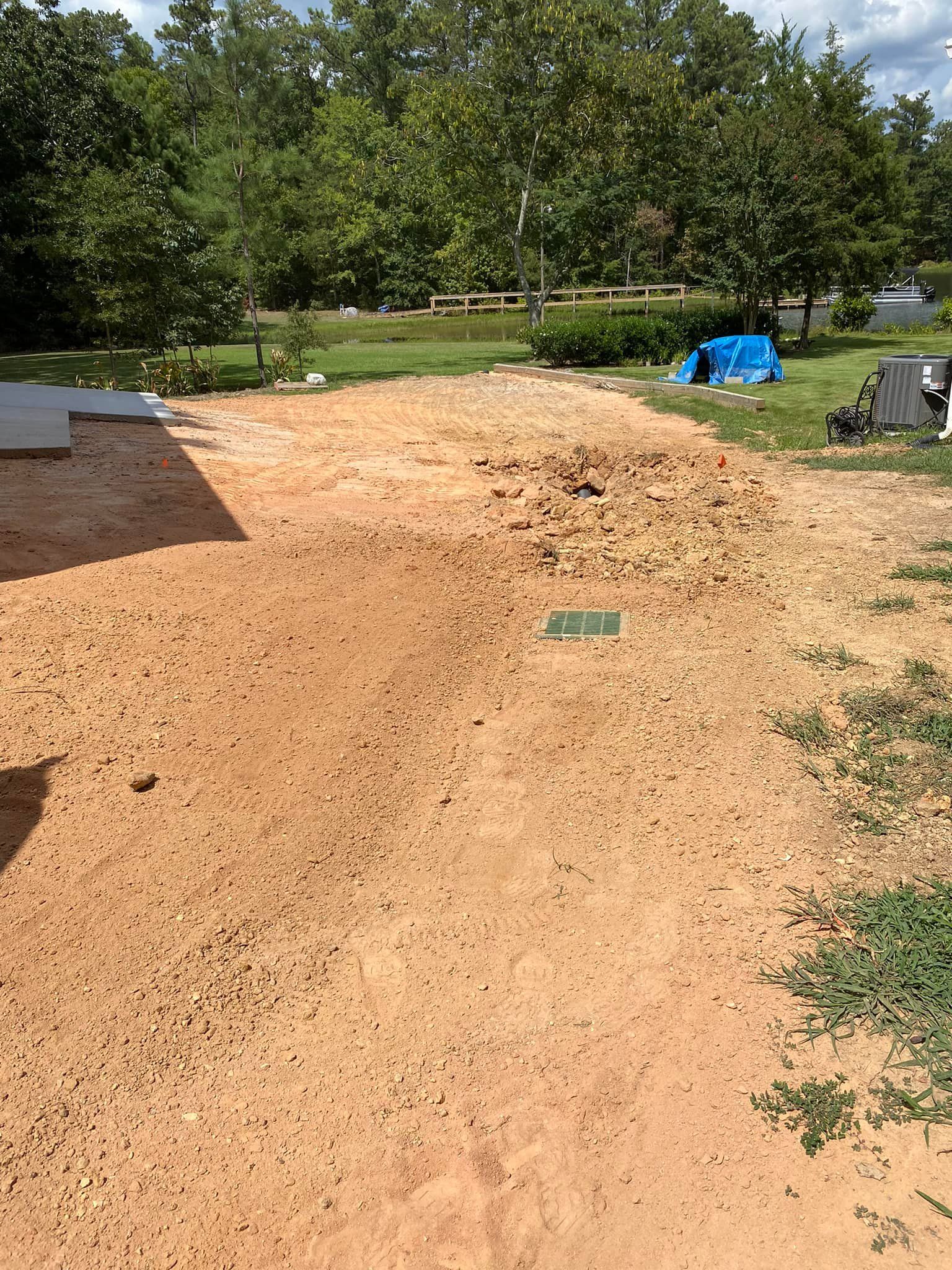 A patch of disturbed, light brown soil with a green utility box, next to a grassy yard with trees and a blue tarp.