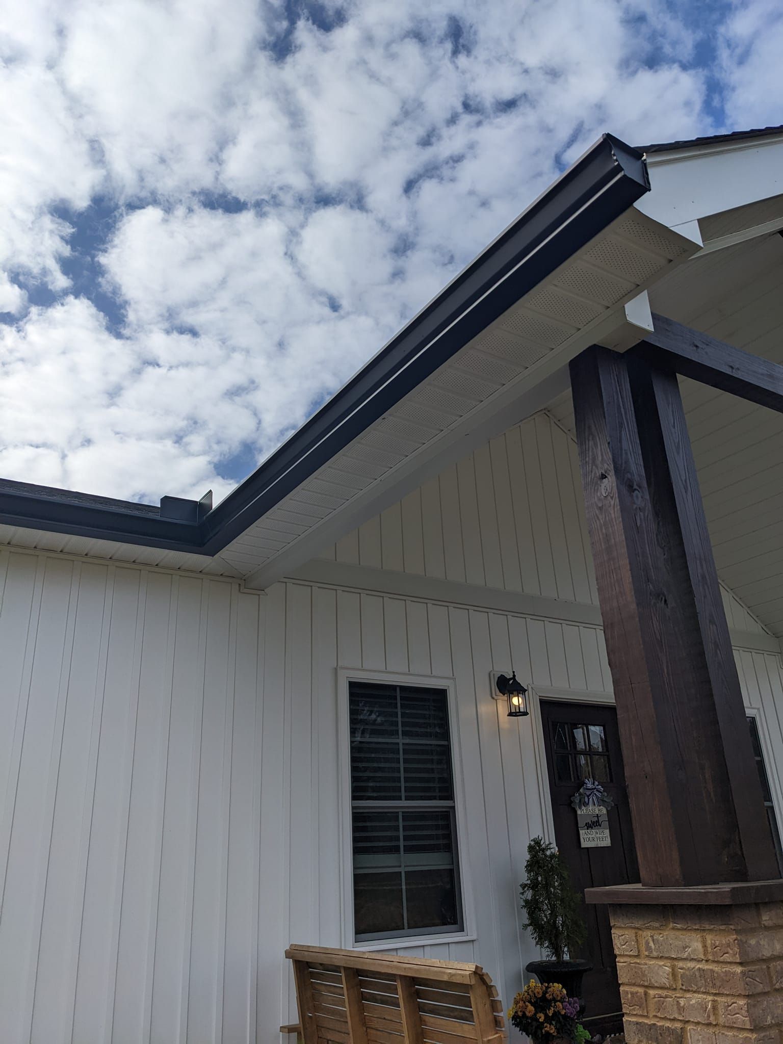 A white house with vertical siding, a dark roofline, a window, and a wooden porch post under a cloudy sky.