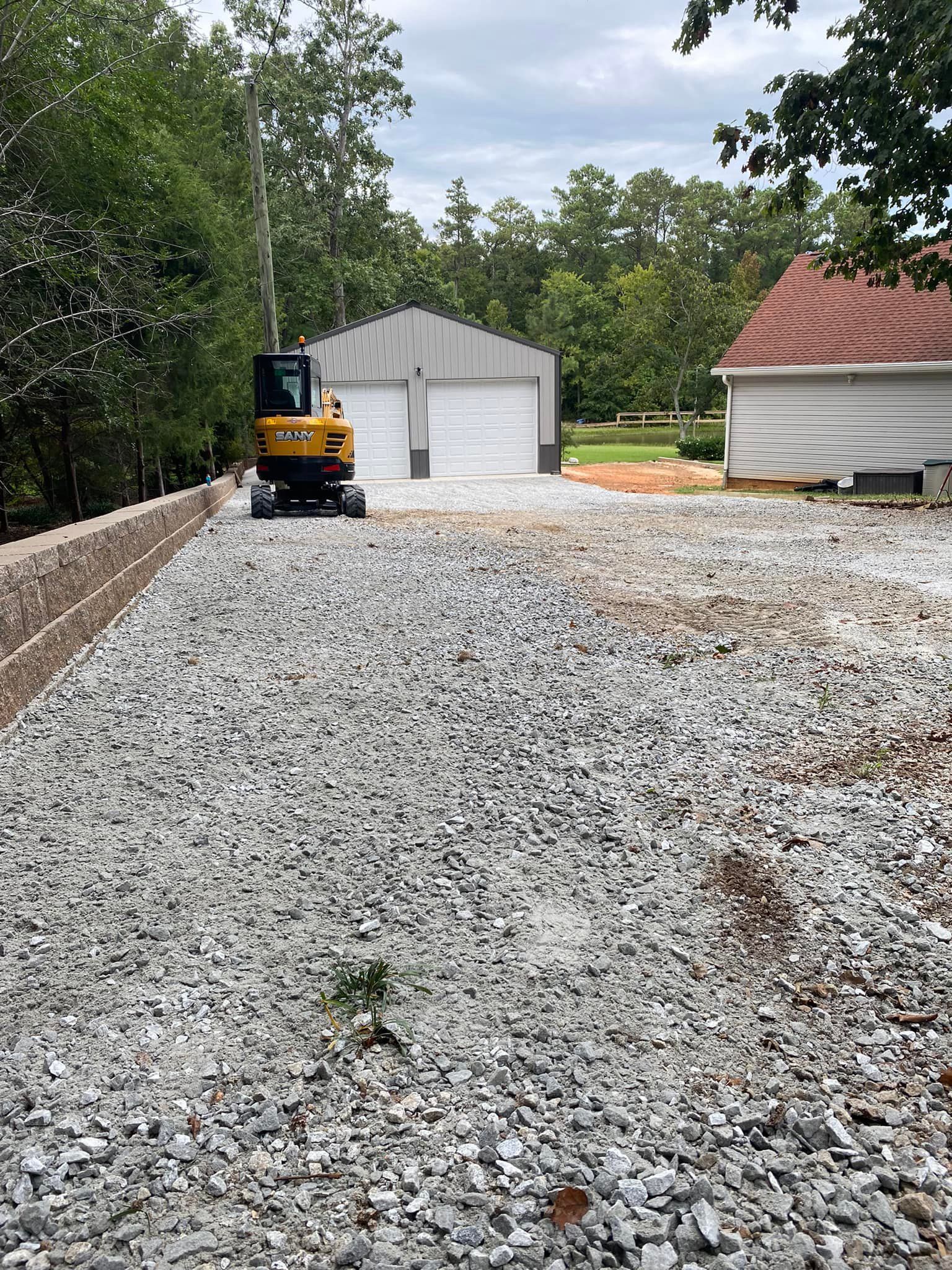 A yellow mini excavator sits on a gravel driveway between a brick retaining wall, a detached garage, and a house.
