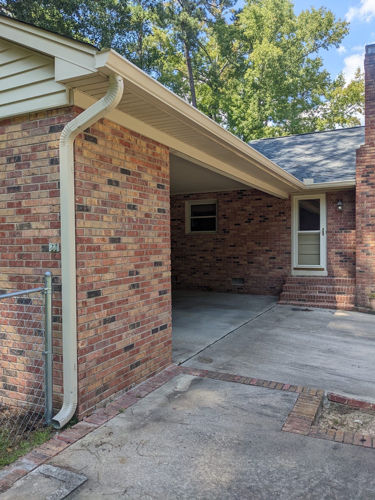 A brick home exterior with a covered carport, featuring a beige downspout and a screen door, set against leafy trees.