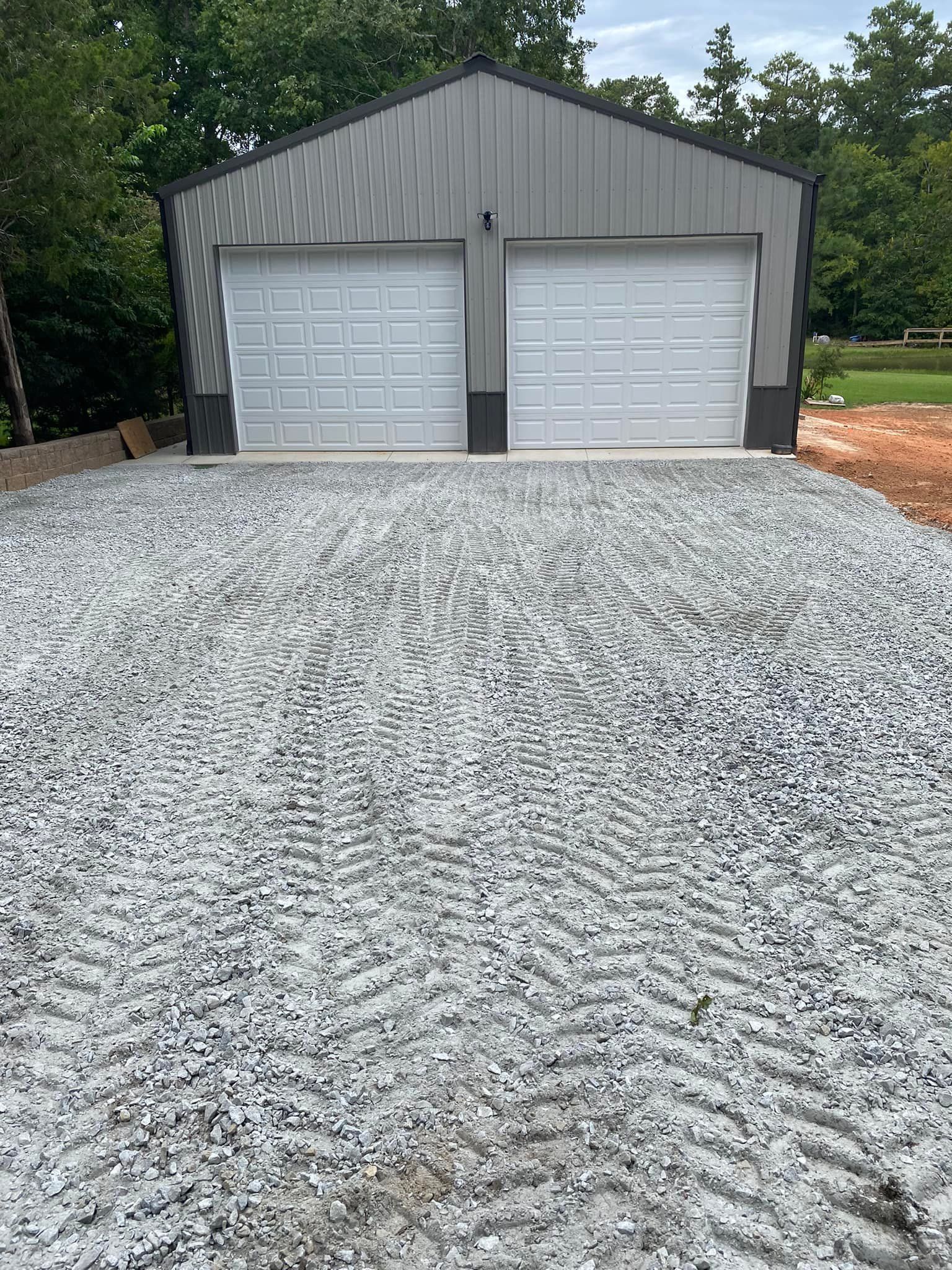 A gray metal two-car garage with white doors sits at the end of a freshly graveled driveway.