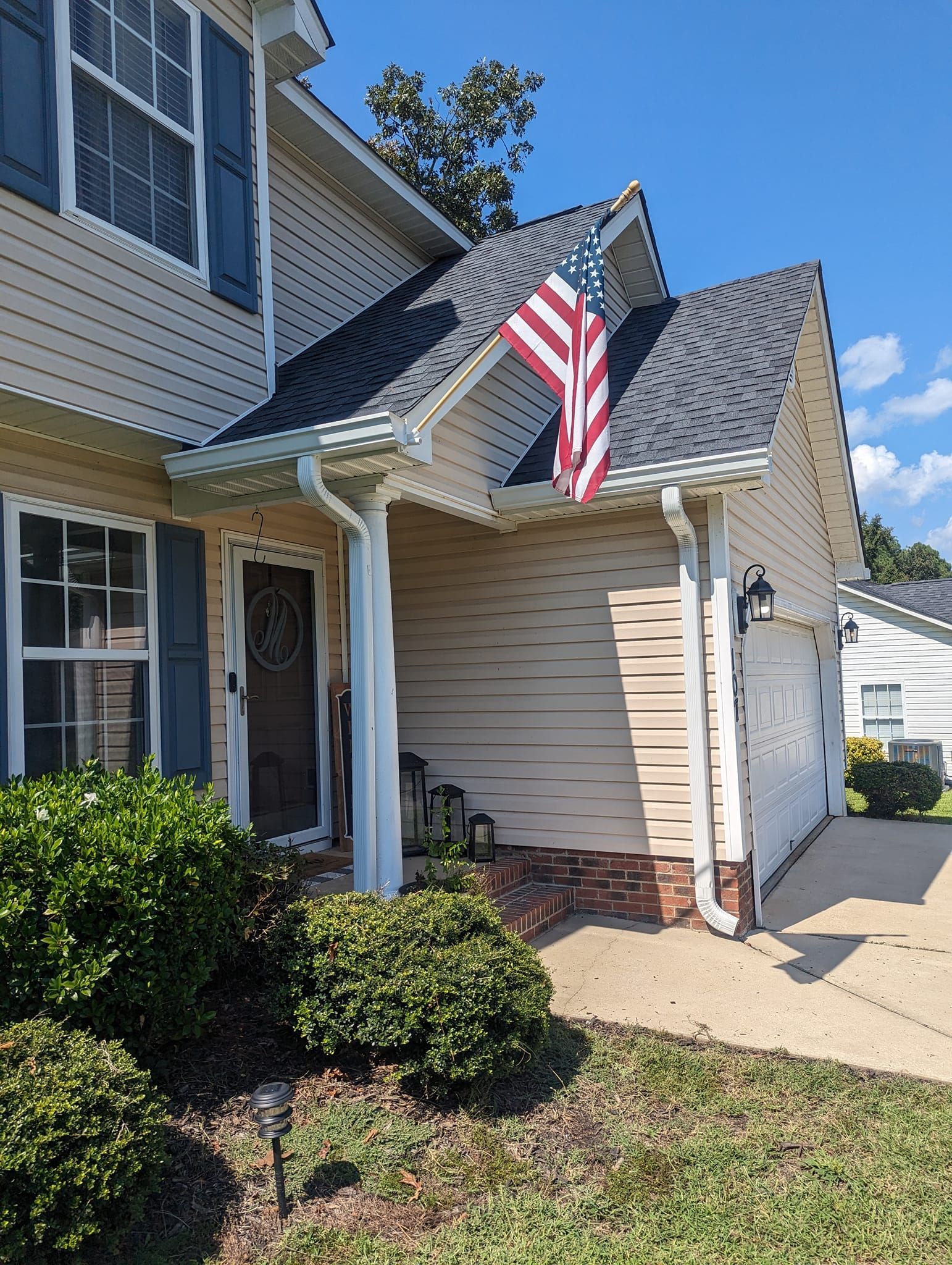 A beige two-story suburban house with blue shutters, a brown front door, and an American flag displayed near the garage.