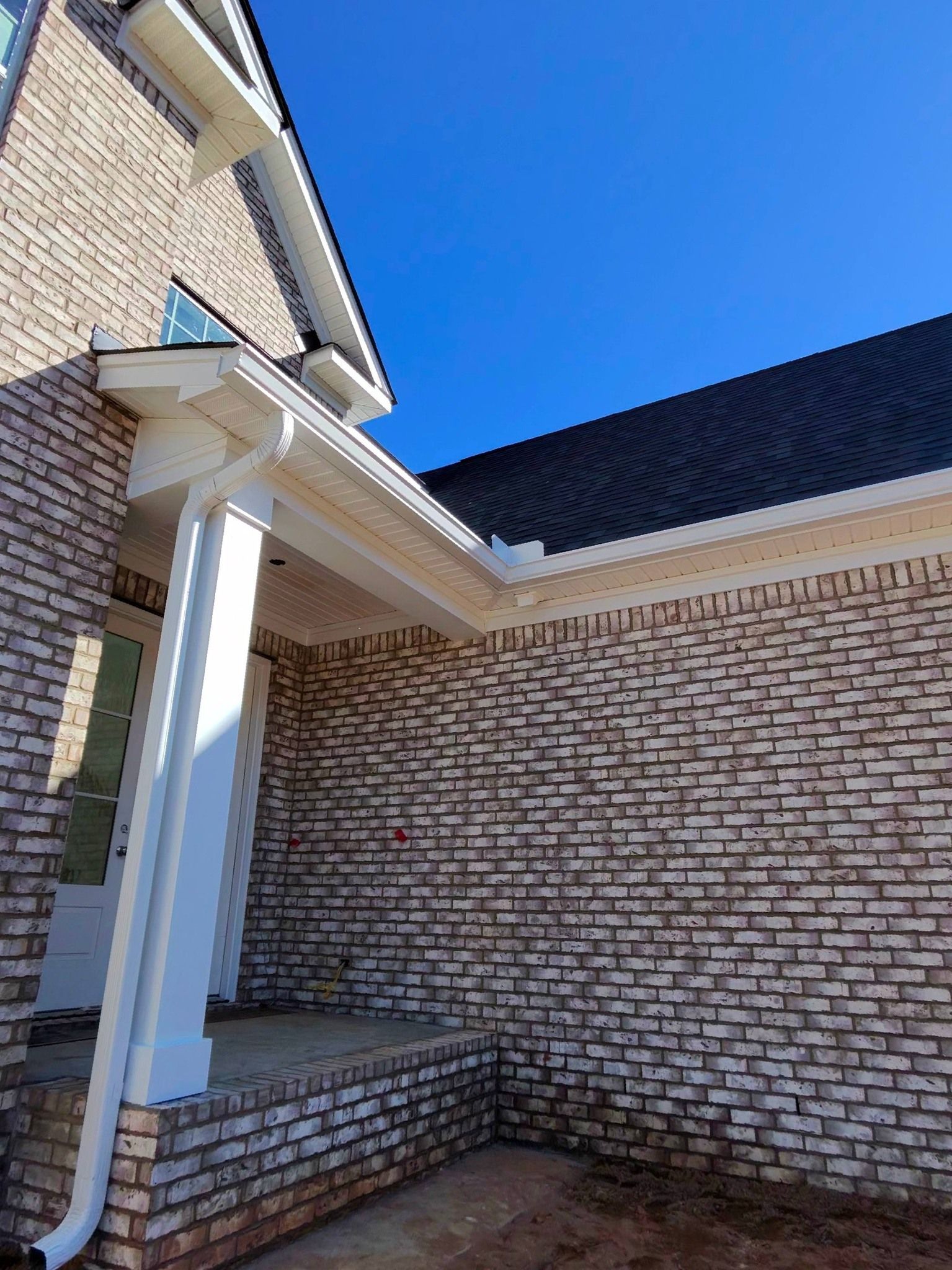 Low-angle view of a modern house exterior with light-colored brick, white columns, and a dark shingled roof under blue sky.