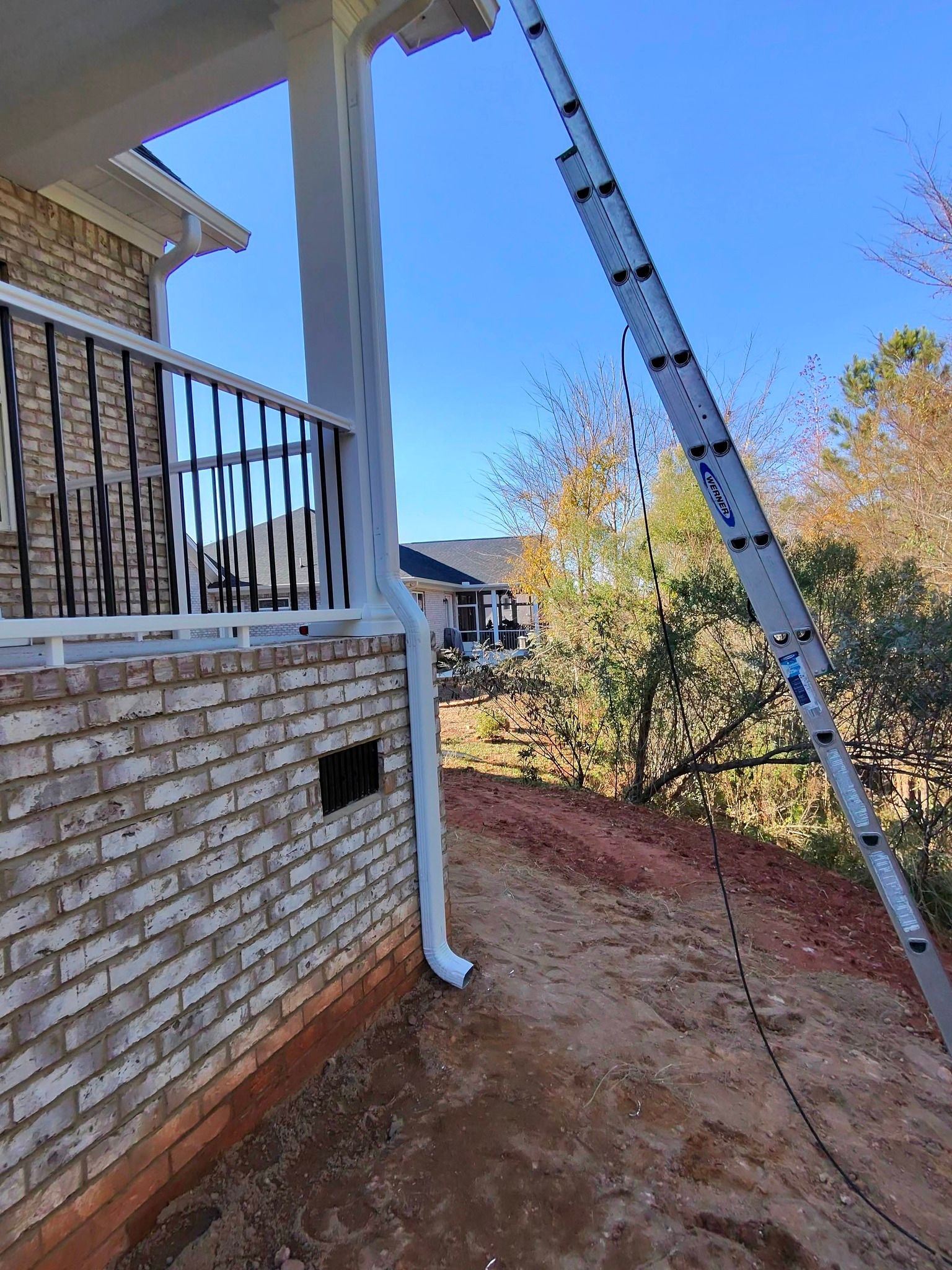 An extension ladder leans against the corner of a brick porch, next to a white downspout on a sunny day.
