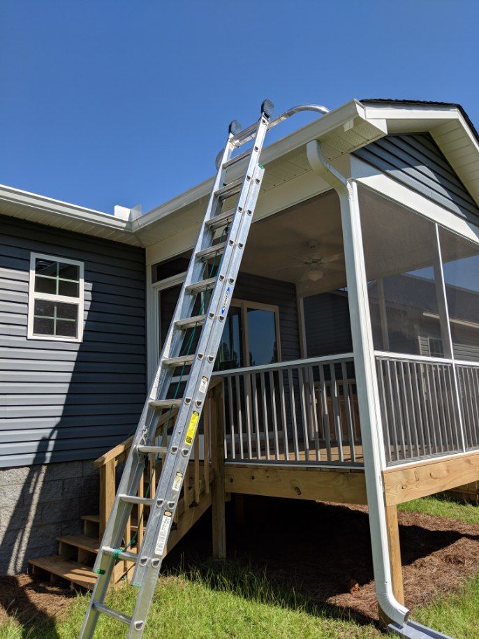 An aluminum extension ladder is leaned against the roof edge of a house with gray siding and an attached screened-in porch.