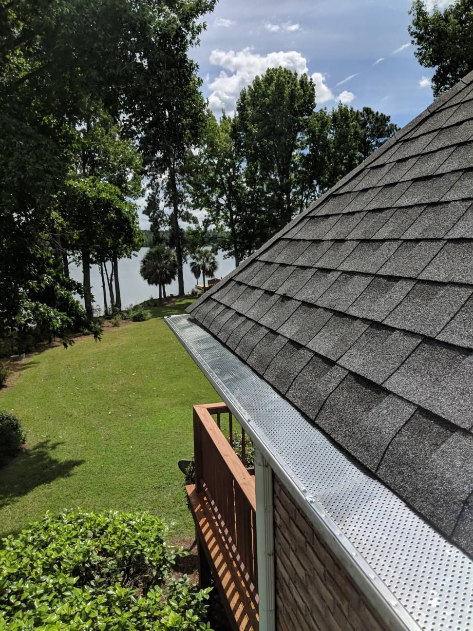 A silver gutter guard installed along the edge of a dark grey shingled roof, overlooking a grassy yard and a lake.