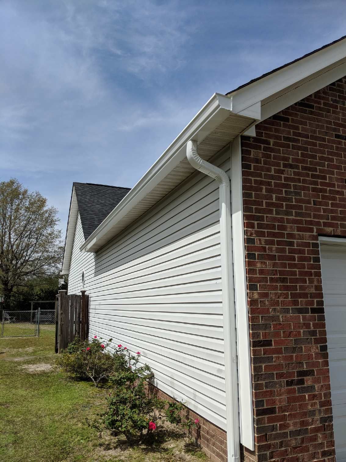 Side view of a house with white horizontal siding, a brick front, white gutter system, and a small rose bush in the yard.