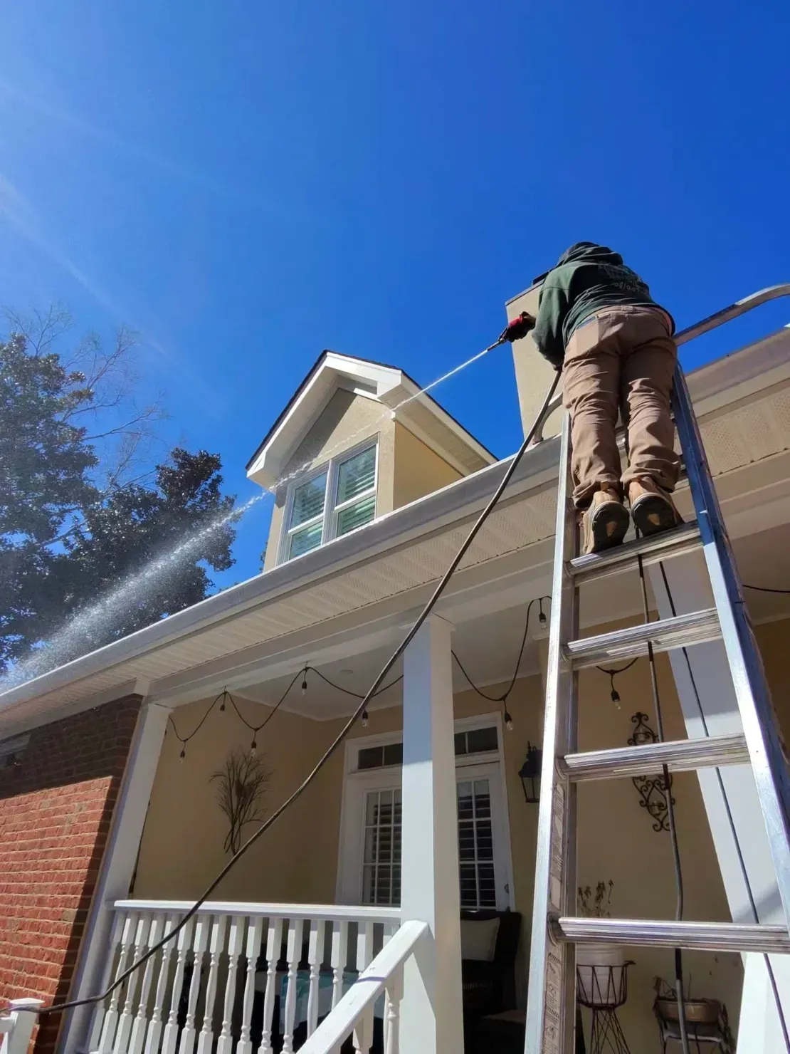 A worker on a ladder power washes the exterior of a cream-colored house under a clear blue sky.