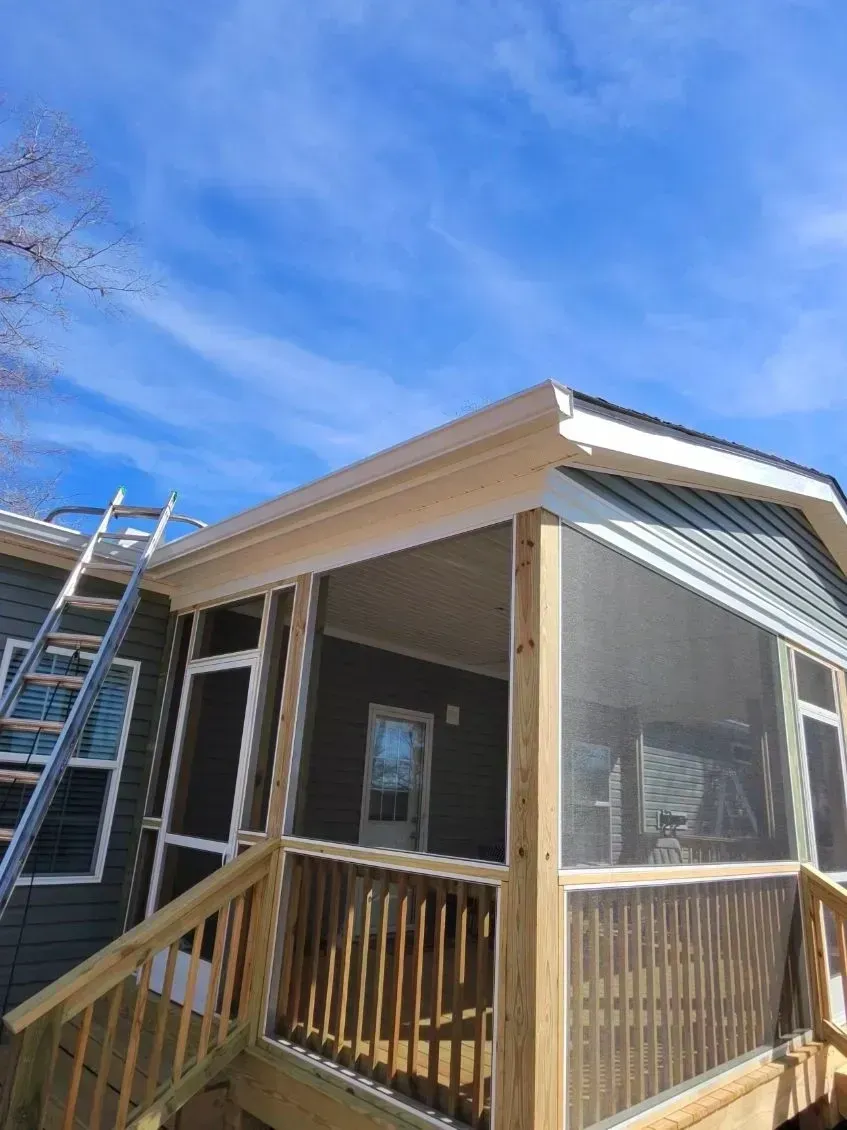 A newly constructed wooden screened-in porch attached to a dark gray house under a clear blue sky, with a ladder visible.