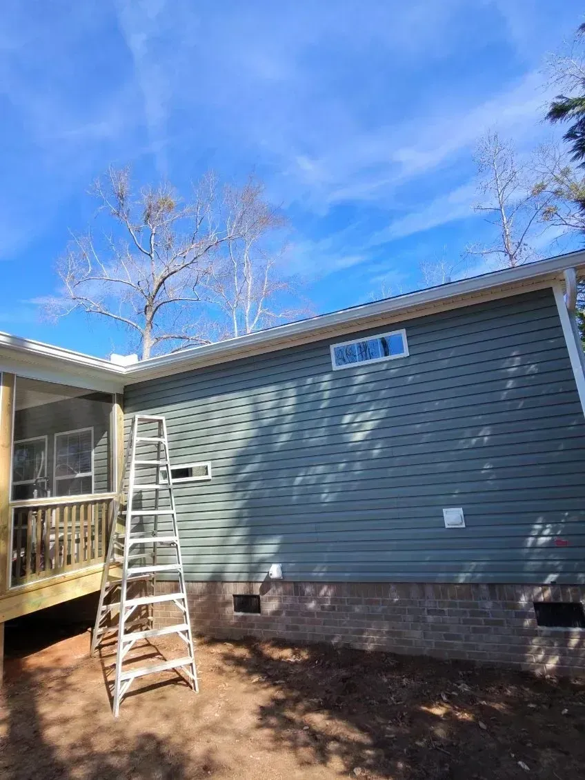 A metal ladder stands against a green-sided house with a stone foundation under a bright blue sky.