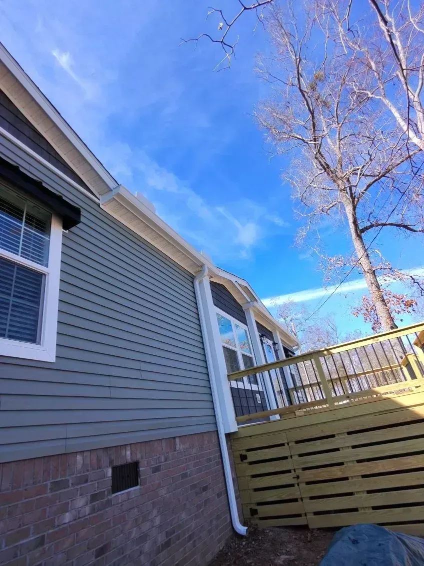 A low-angle view of a house with grey siding, brick foundation, a white gutter system, and an attached wooden deck.