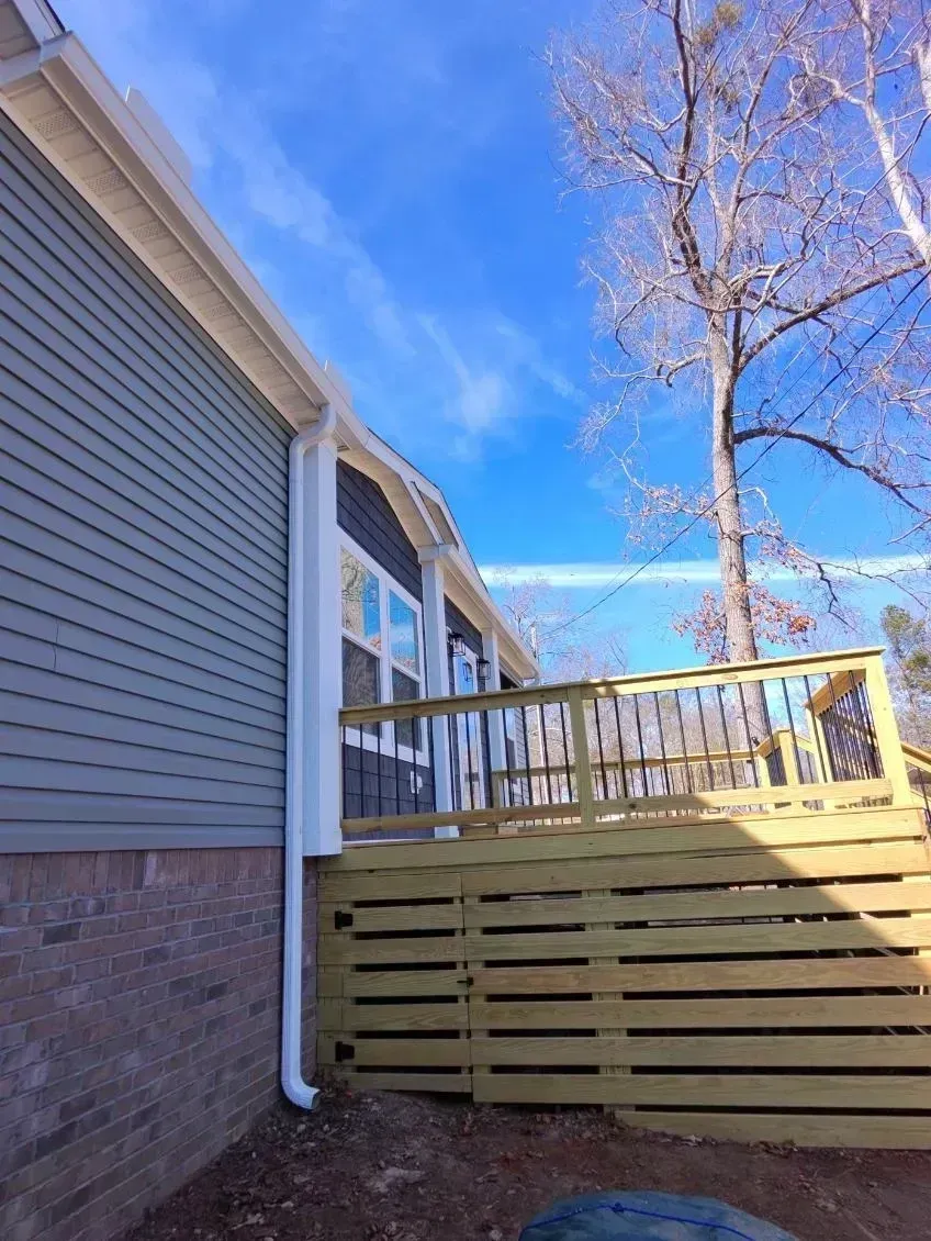 A side view of a house with gray siding, a brick foundation, a white gutter downspout, and a wooden deck.