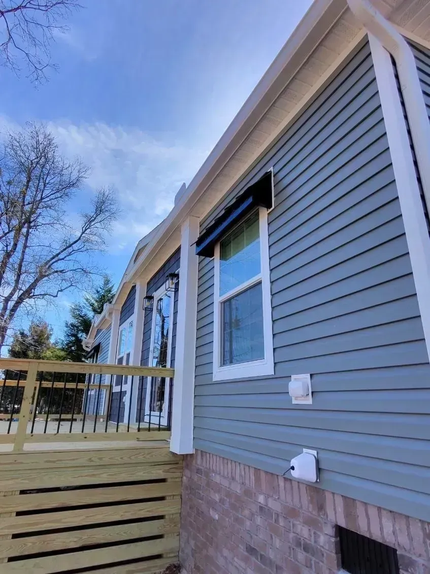 A low-angle view of a blue-sided house with a brick foundation, featuring a wooden deck and a black-framed window.