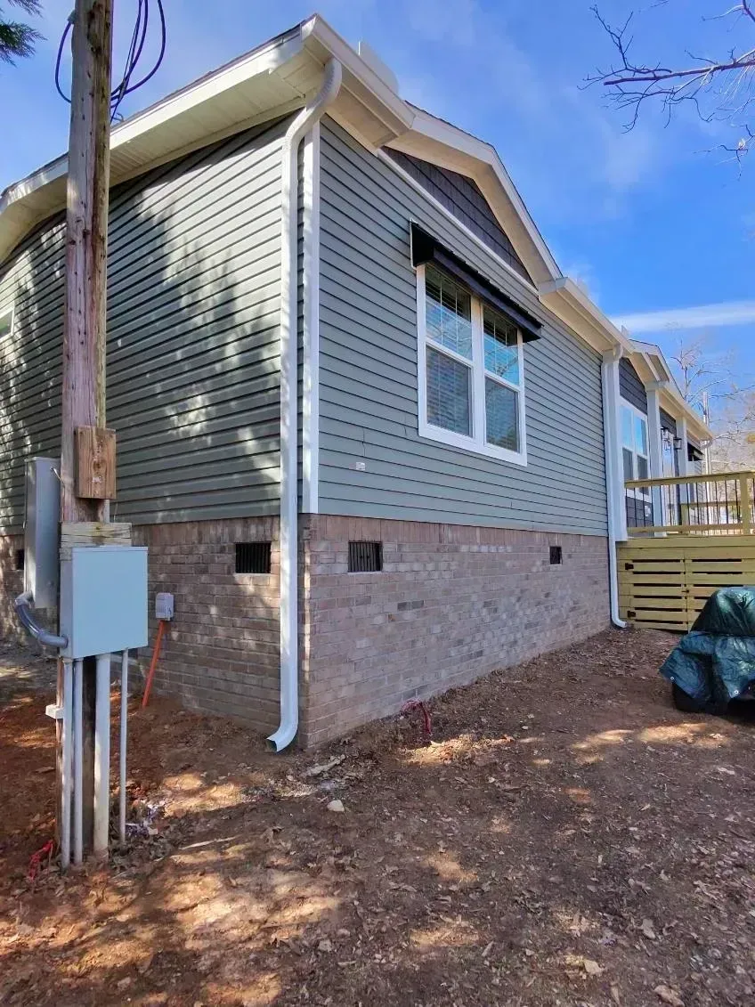 Light green, vinyl-sided house with a brick foundation and white downspouts, viewed from the side on a sunny day.
