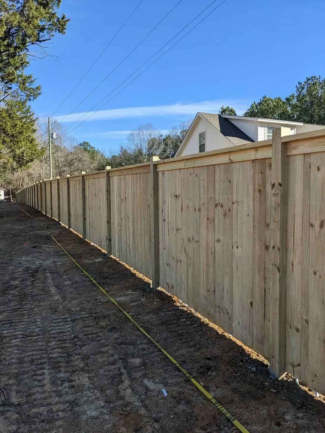 A newly installed wooden privacy fence extends along a dirt path under a clear blue sky.