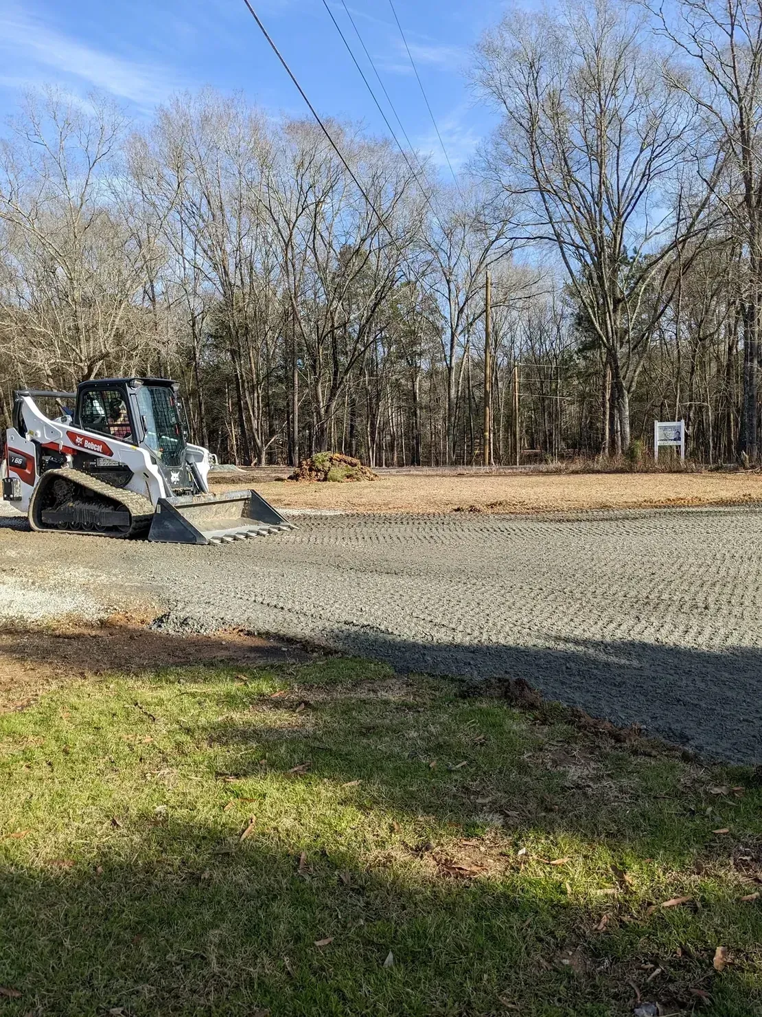 A white Bobcat skid-steer loader parked on a freshly graveled area next to a grassy lawn and a wooded backdrop.
