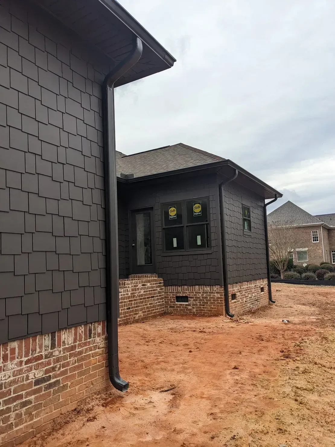 A partially constructed house with dark, square-patterned siding, brick foundations, and black gutters against a cloudy sky.