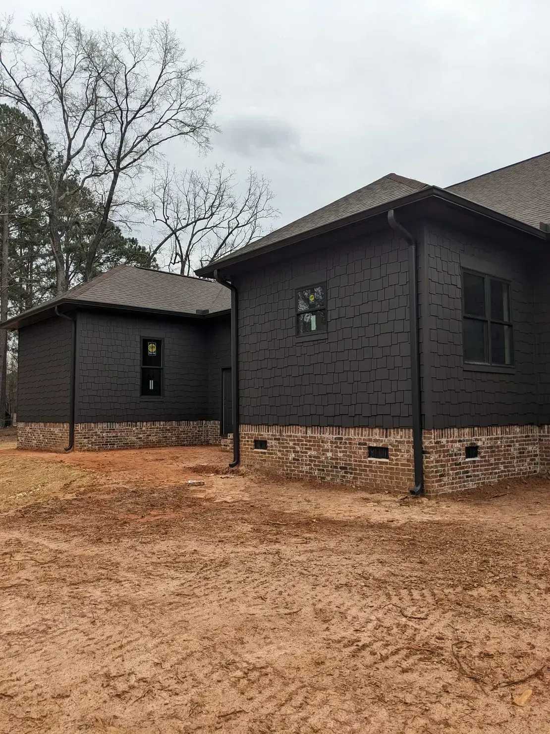 Dark textured siding homes with stone foundations sit on a dirt lot under an overcast sky.