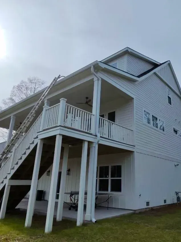 A white, elevated multi-story house with a large wooden staircase and a covered porch area at ground level.