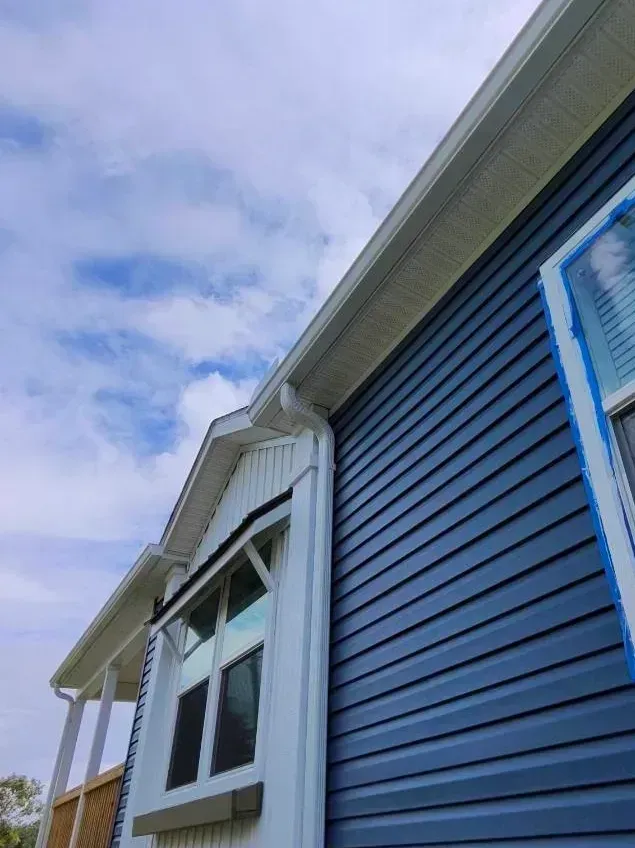 A low-angle view of a blue-sided house exterior with a white-framed window, porch area, and a white gutter against a sky.