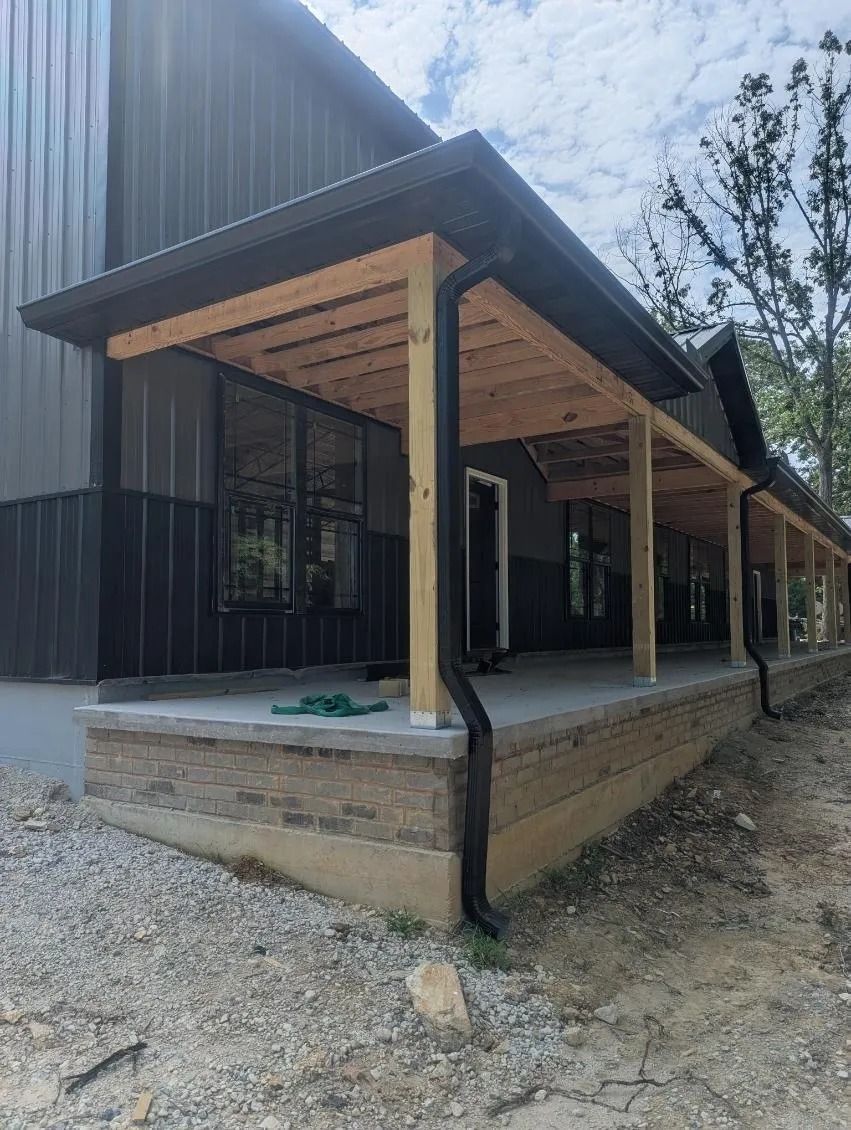 A metal-sided building with a long, unfinished wooden porch overhang and a black downspout over a gravel-covered yard.