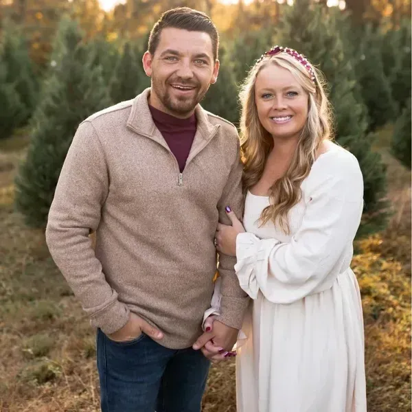 A couple stands in a Christmas tree farm, smiling and holding hands in an outdoor, sunlit setting.