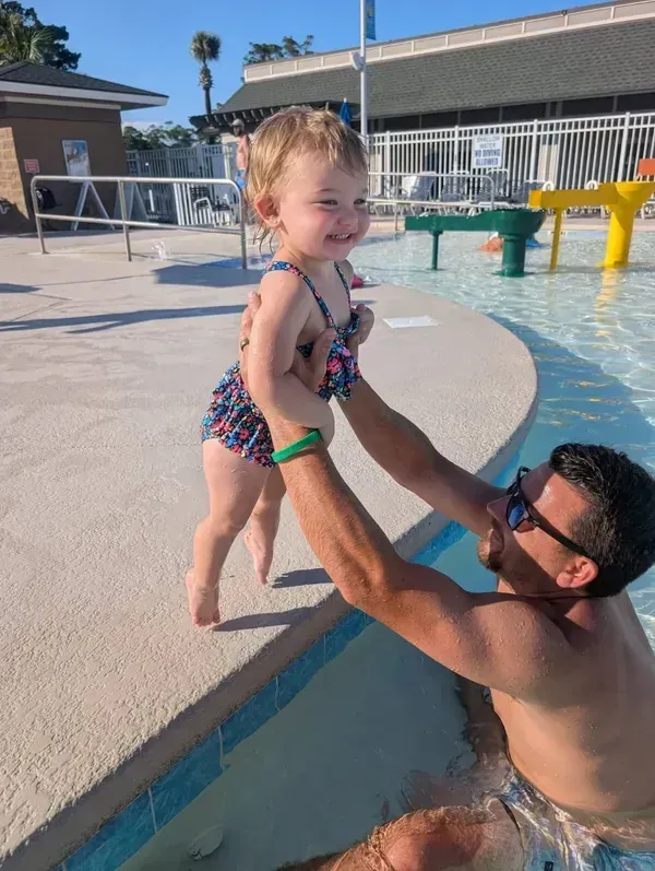 A person in a swimming pool lifts a toddler in a patterned swimsuit out of the water toward the pool deck.