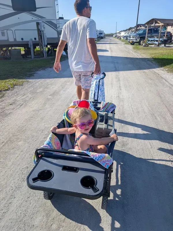 A person pulls a child in a wagon along a dirt path lined with parked vehicles and campers on a sunny day.