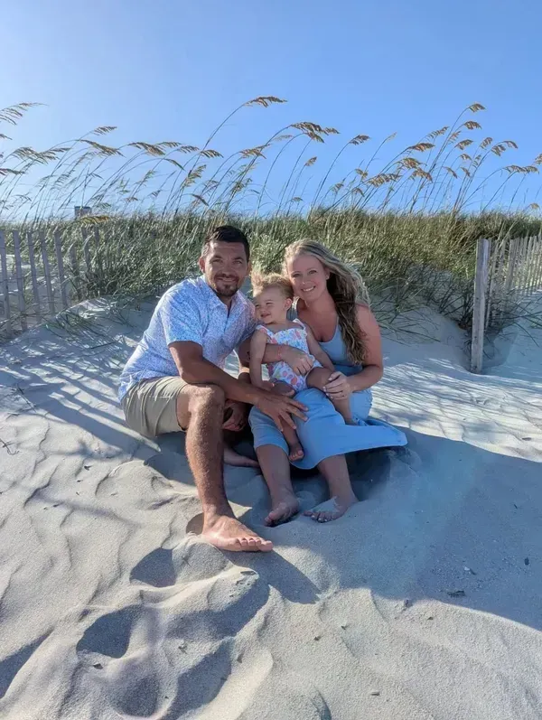 A family of three sitting on sandy dunes in front of beach grass under a clear blue sky.