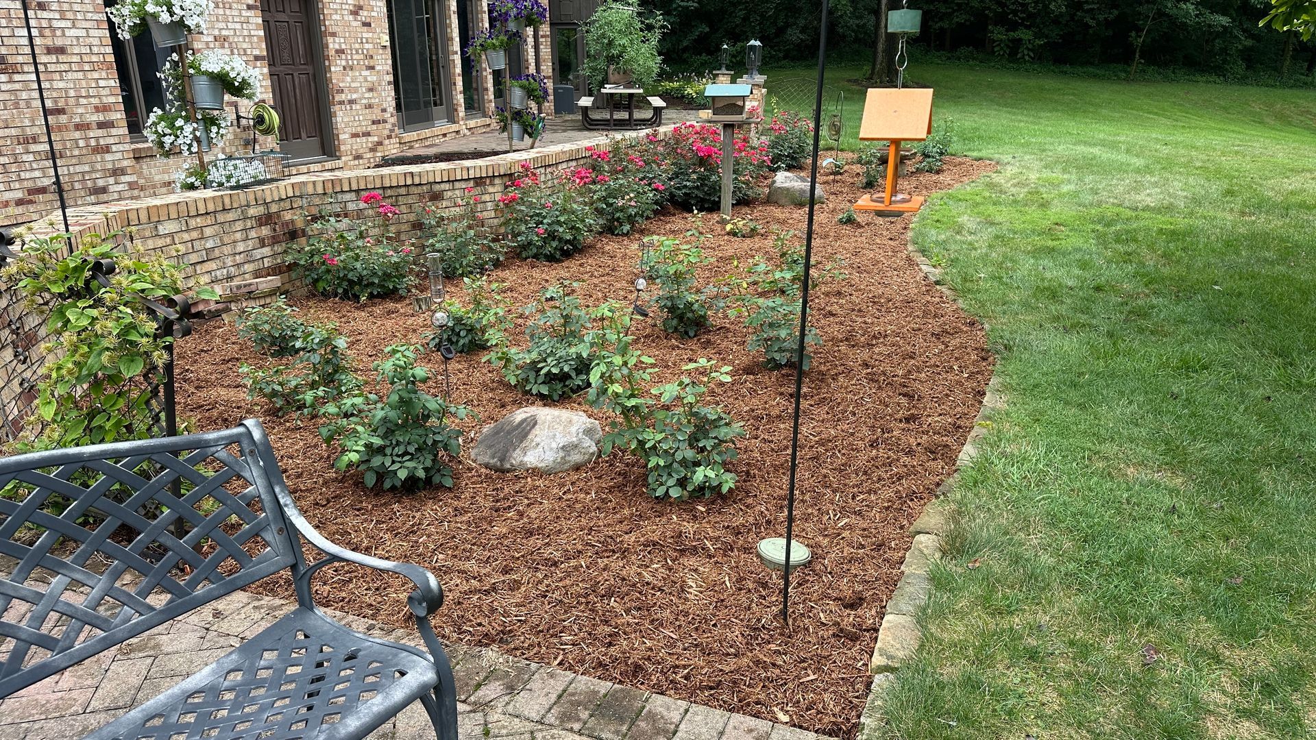 Mulched garden bed with young plants, stone edging, lawn, and a bench beside a brick house