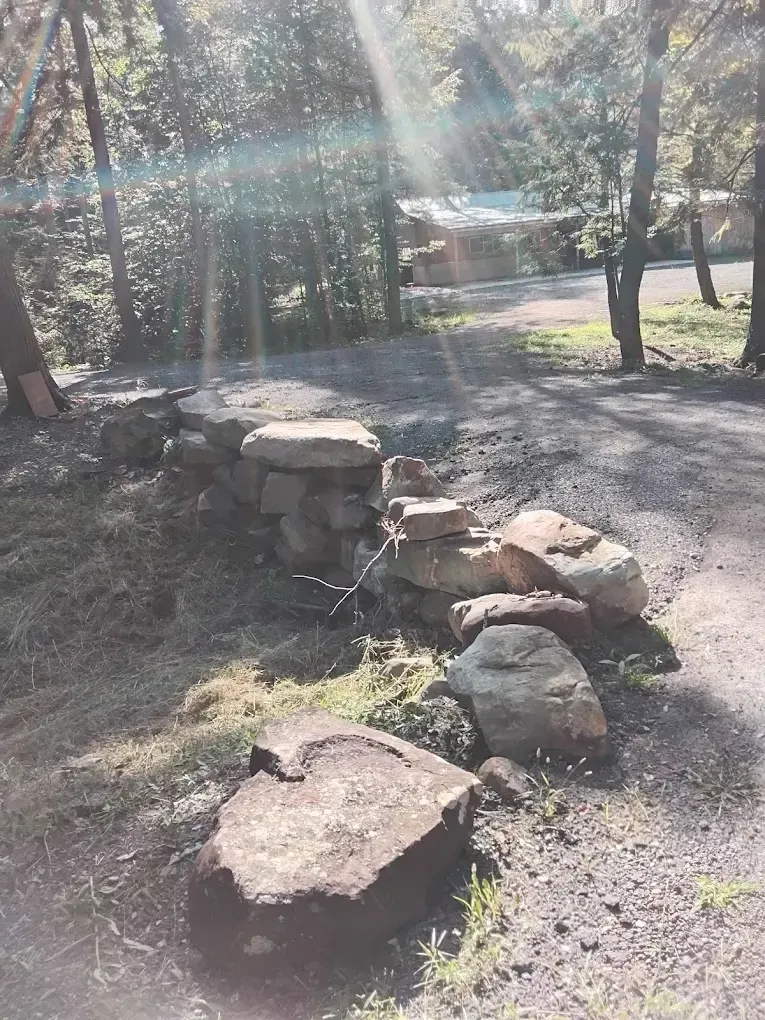 Stone wall along a gravel path in a wooded area, with a cabin visible in the background.