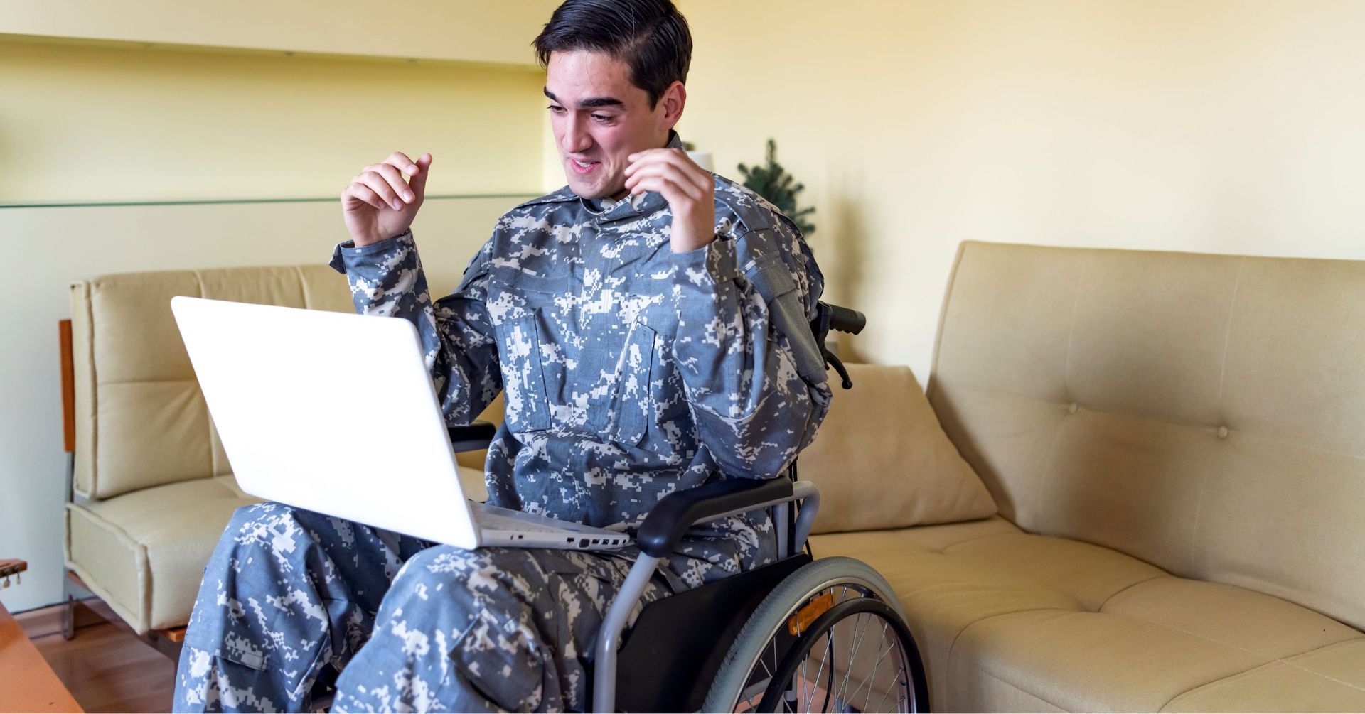 A young male veteran in camouflage uniform sitting in a wheelchair and reacting with excitement while looking at a white laptop on his lap.