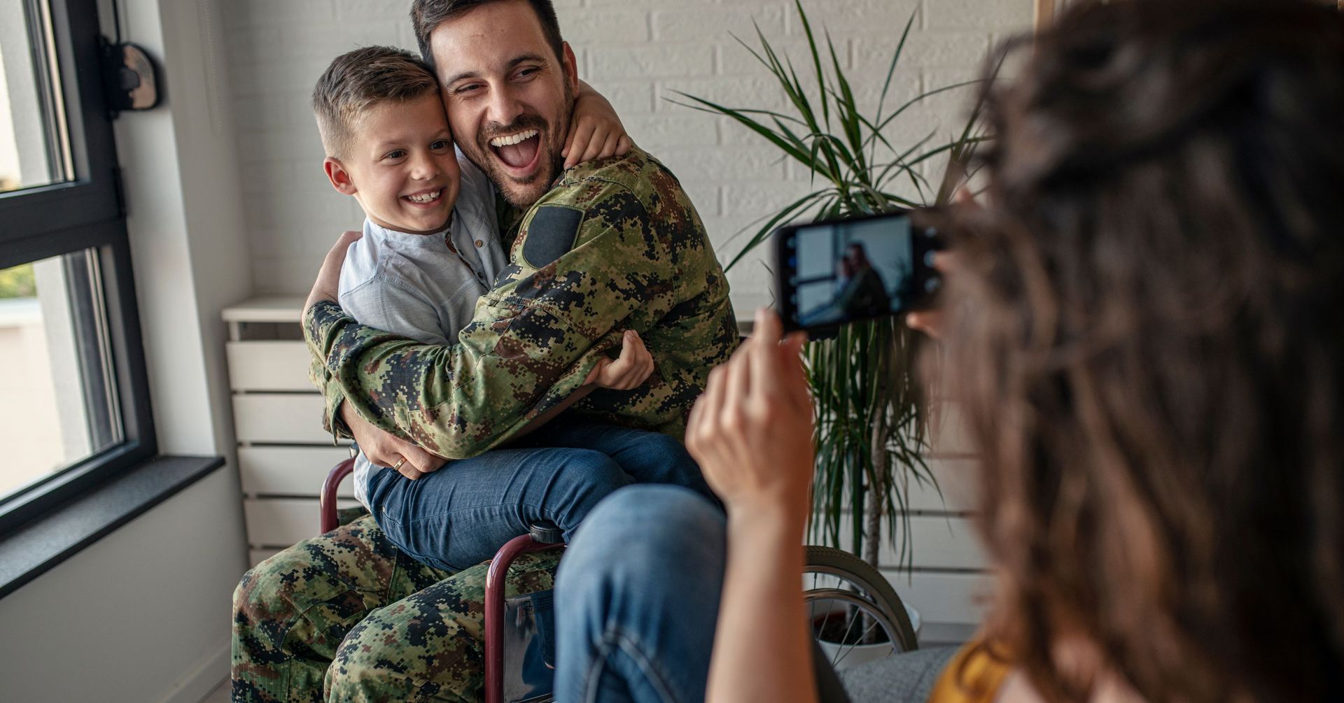 A male veteran in a wheelchair laughing and hugging a young boy while a woman in the foreground takes a photo with a smartphone.